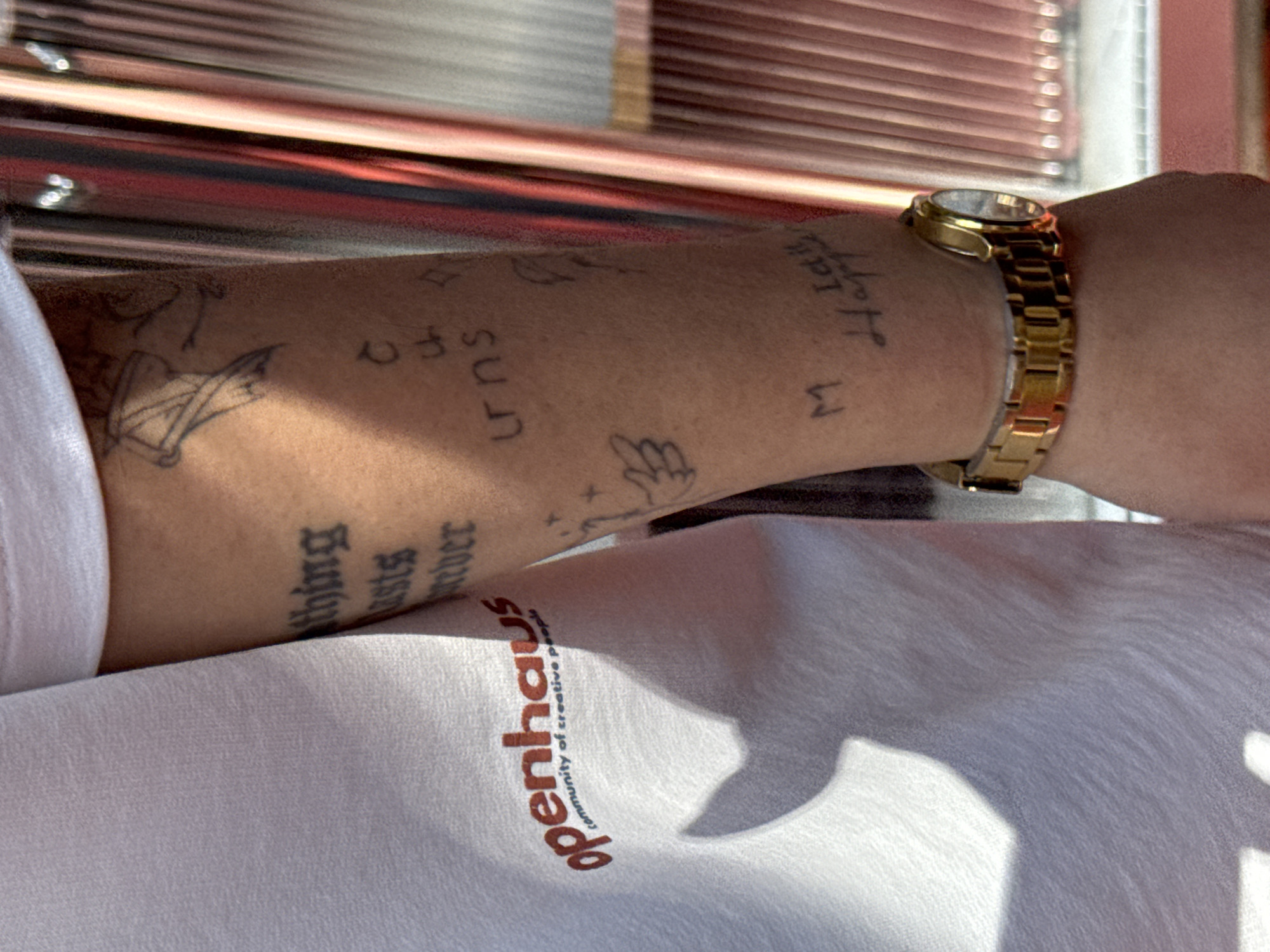 An arm with multiple tattoos is visible, adorned with a gold watch. The tattoos include the word "thing", the word "lasts", the word "never", the word "turns", a small wing and a small star. The writing "M Let's Happy" is also tattooed. A portion of a white shirt with the text "openhaus community of creative people" is also visible. The background is a reddish metal. There is no explicit information to determine if the scene is in Yerevan, Armenia.