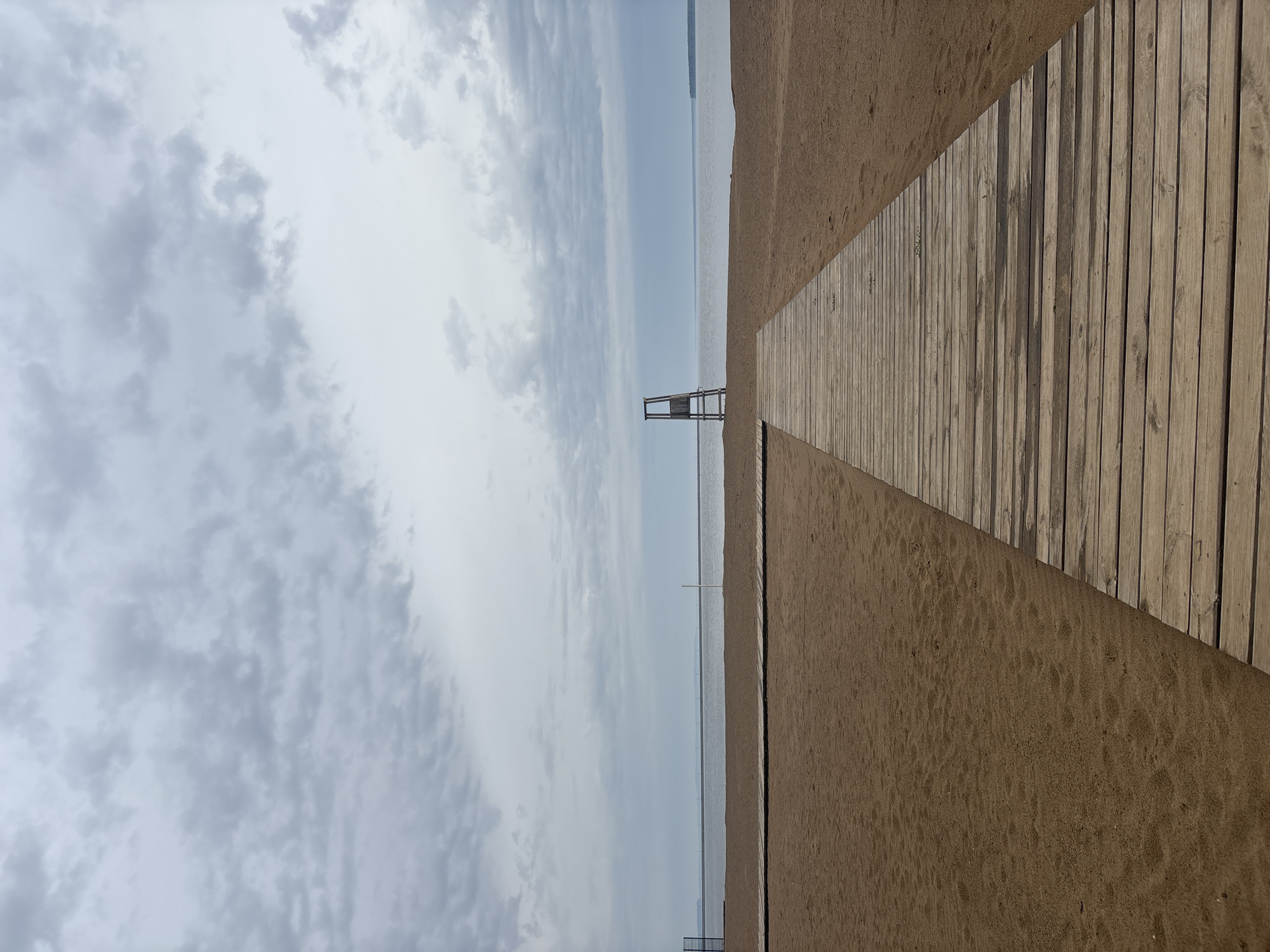 The image shows a beach scene in Chaikovsky, Russia. A wooden boardwalk stretches from the bottom right corner towards the center of the image, leading to the beach and water. Sand is visible to the left of the boardwalk. A lifeguard tower is situated further out towards the water. The sky is overcast with clouds. The water is calm and reflects the sky.