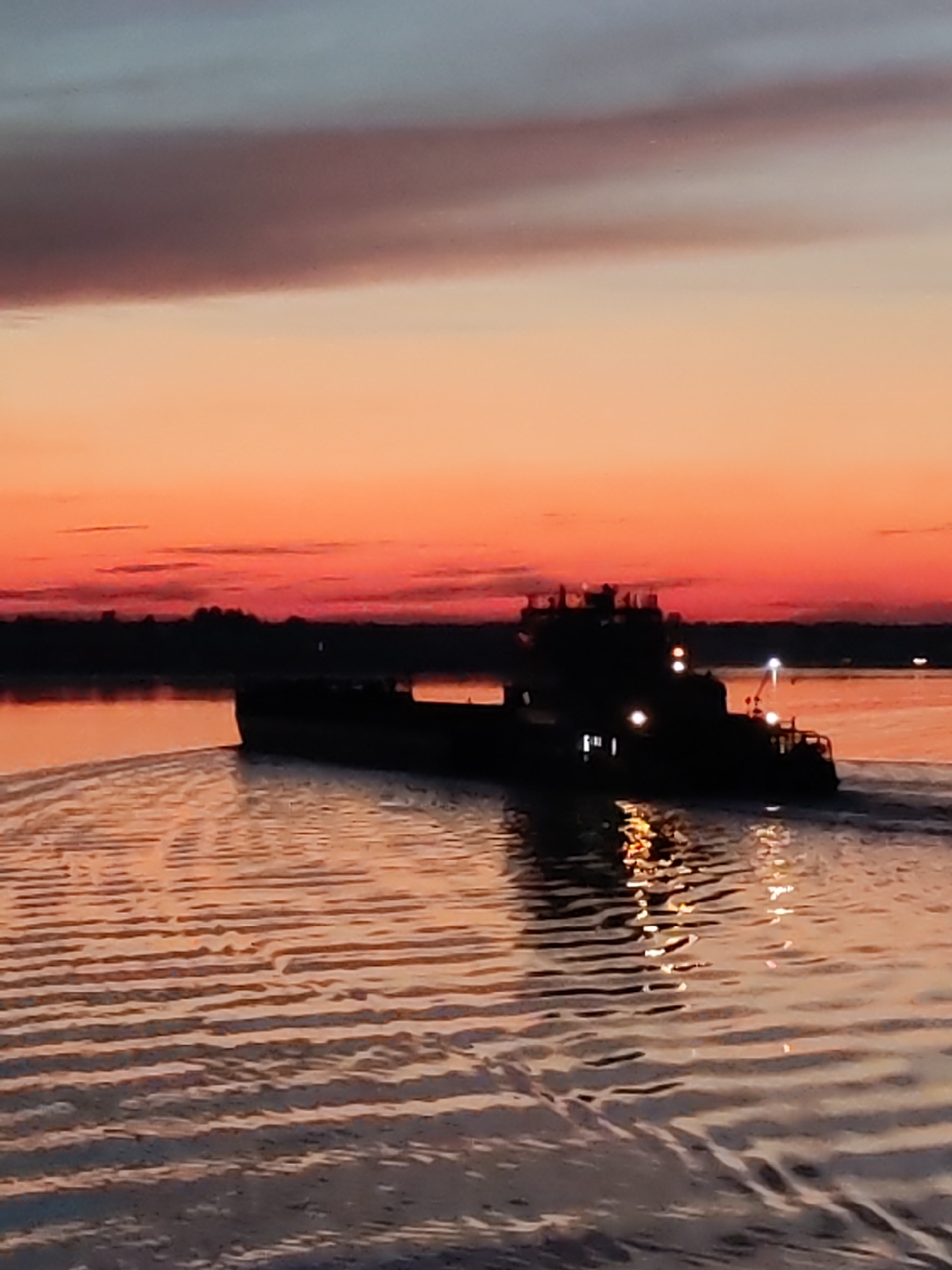 An over-the-shoulder drone shot captures a silhouette of a ship navigating a body of water in Babnya, Russia. The sky above displays a gradient of colors, transitioning from a dusky blue at the top to a vibrant orange and pink sunset near the horizon. The water's surface is rippled, reflecting the warm hues of the sky and the artificial lights of the ship. The ship itself is a dark silhouette, with visible lights on its structure, indicating it is a powered vessel. The background features a dark, tree-lined silhouette of the shore, further emphasizing the sunset's glow. The overall impression is one of tranquility and the end of a day, with the ship a lone traveler on the calm waters.