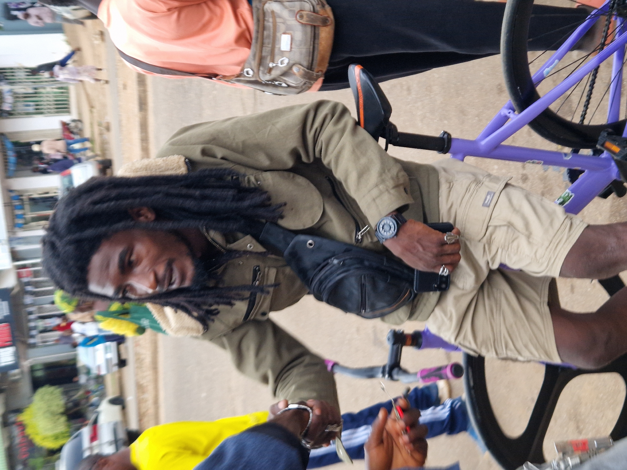 In an outdoor urban setting, a Black man with dreadlocks, wearing a green jacket with a shearling collar, khaki shorts, and a black crossbody bag, sits on a purple bicycle. He is holding a set of keys and a smartphone. To his left, another person in a blue shirt and pants is partially visible, as is a man wearing a yellow shirt. Another person in an orange shirt stands to the man's right, with a beige purse slung over their shoulder. In the background, several cars and a building with storefronts are visible, suggesting a street or commercial area. The ground is paved with dirt and asphalt.