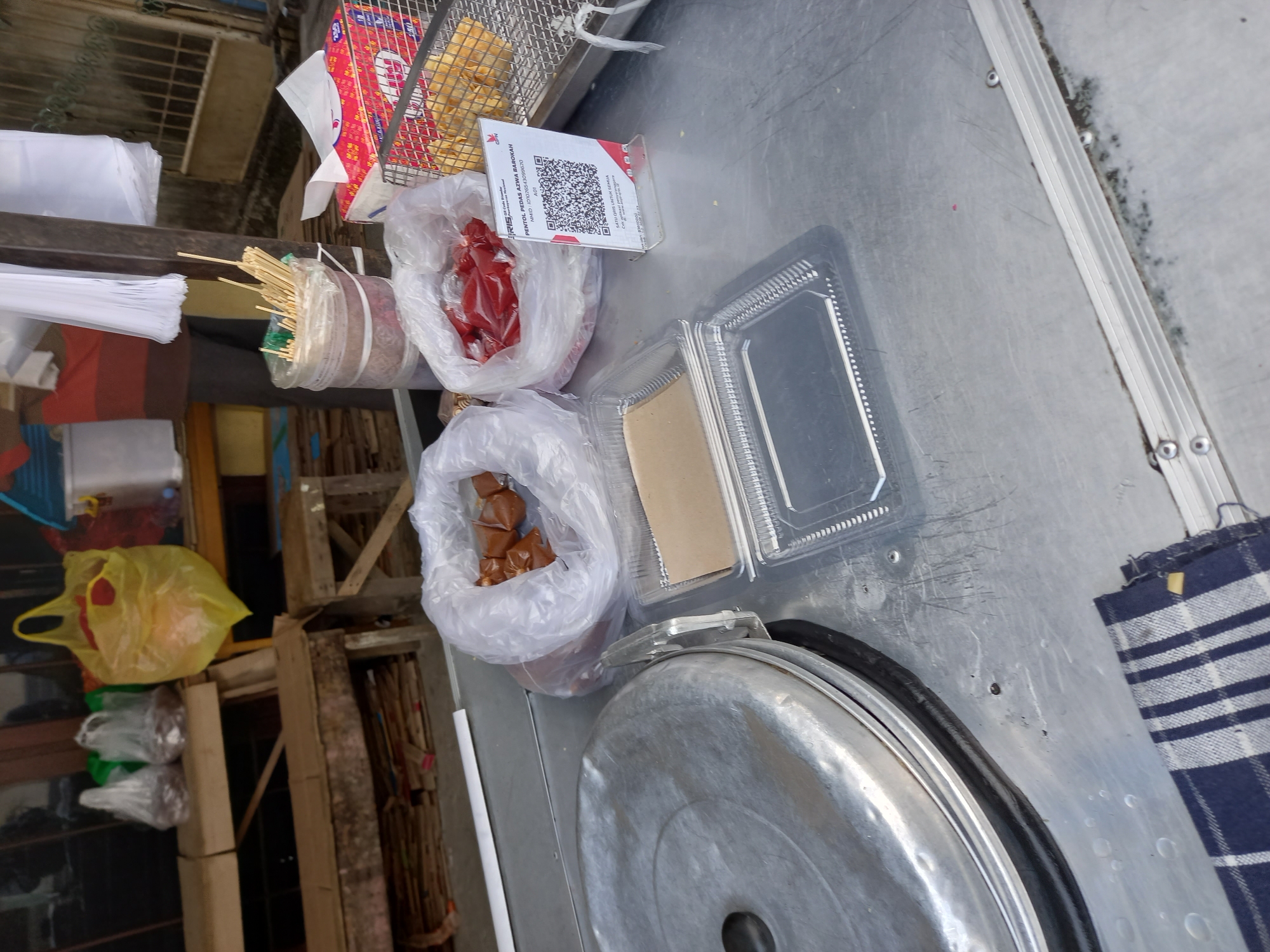 This is a medium-shot, eye-level, outdoor, street-level photograph taken in the city of Samarinda, Indonesia. The scene depicts a food cart with various items displayed on its metal surface. In the foreground, to the left, is a large, lidded metal pot, possibly a steamer. To its right are stacks of clear plastic containers, some empty and some with brown paper liners.
Further back on the cart, there are two plastic bags, one containing small, brownish, sausage-like items, and another filled with a red, sauce-like substance. Behind these bags, a QR code payment stand is visible. To the far right, a metal basket holds fried snacks. The background shows wooden shelves with items in plastic bags, a yellow plastic bag, and some blurred structures and vegetation. A person in a striped shirt is partially visible in the background. The overall impression is of a bustling street food vendor setup.