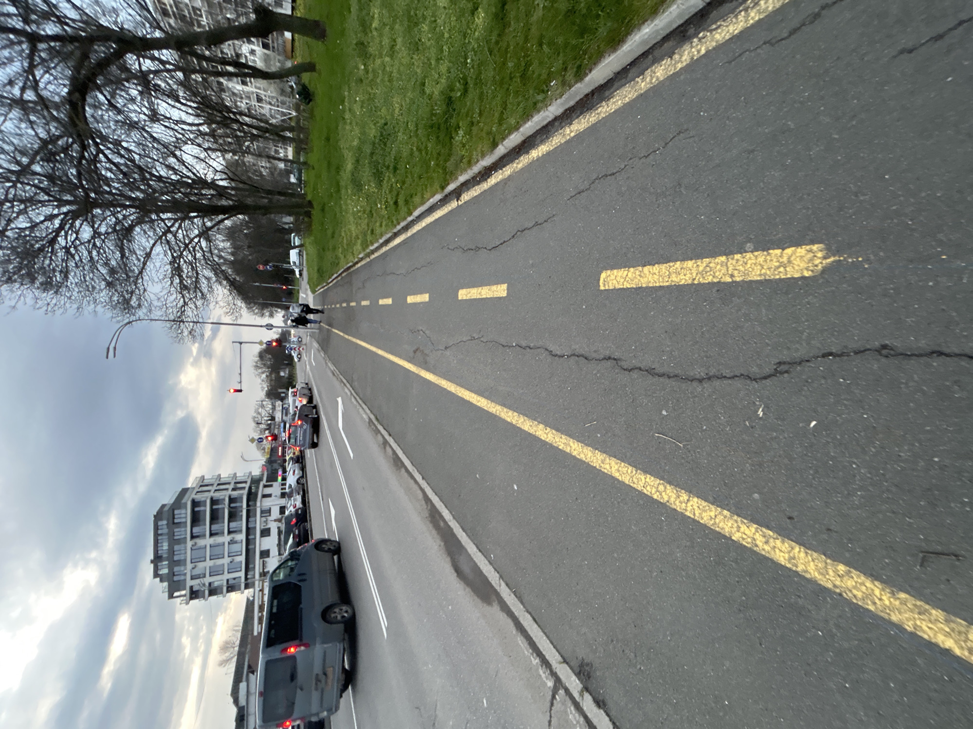 The image shows a section of a street in Burgas, Bulgaria. A dedicated bike path, marked with yellow lines, runs along the right side of the road, separated from the adjacent grassy area by a curb. Cracks are visible on the asphalt surface of the bike path. A line of parked and moving vehicles occupies the roadway to the left of the bike path. A multi-story building with a modern architectural design and many windows is visible on the left. Bare trees line the area adjacent to the grassy section. Traffic lights are visible. The sky is overcast. Two people are walking further down the bike path.