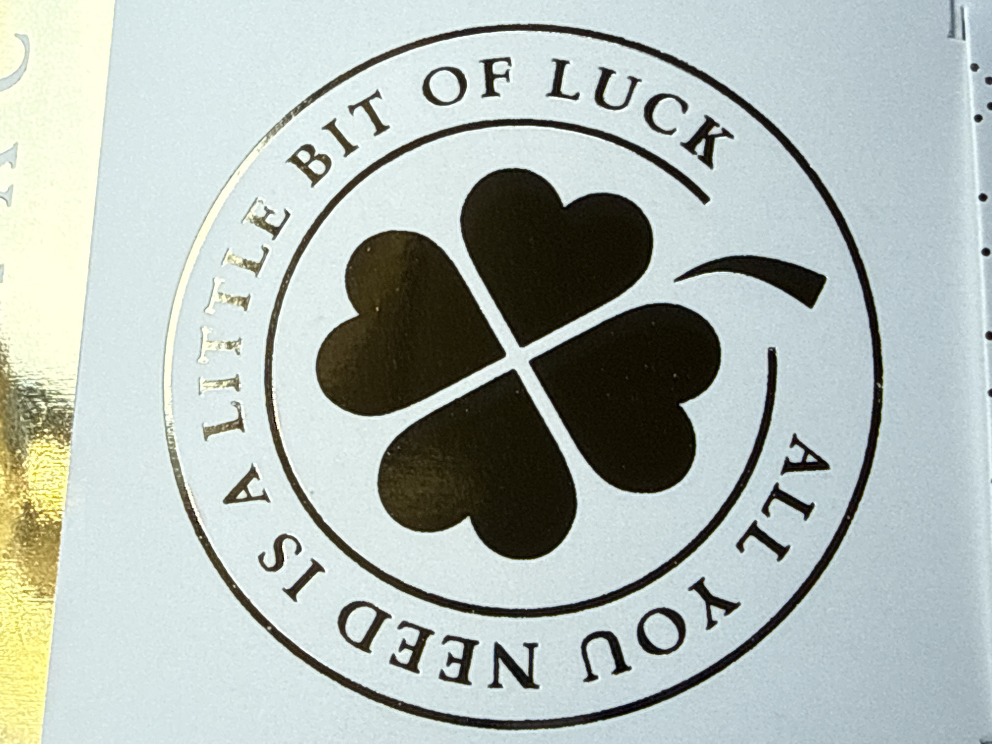 The image shows a gold foil embossed circular design with the text "A LITTLE BIT OF LUCK ALL YOU NEED IS" surrounding a four-leaf clover. The background is a light, pale blue/white.
