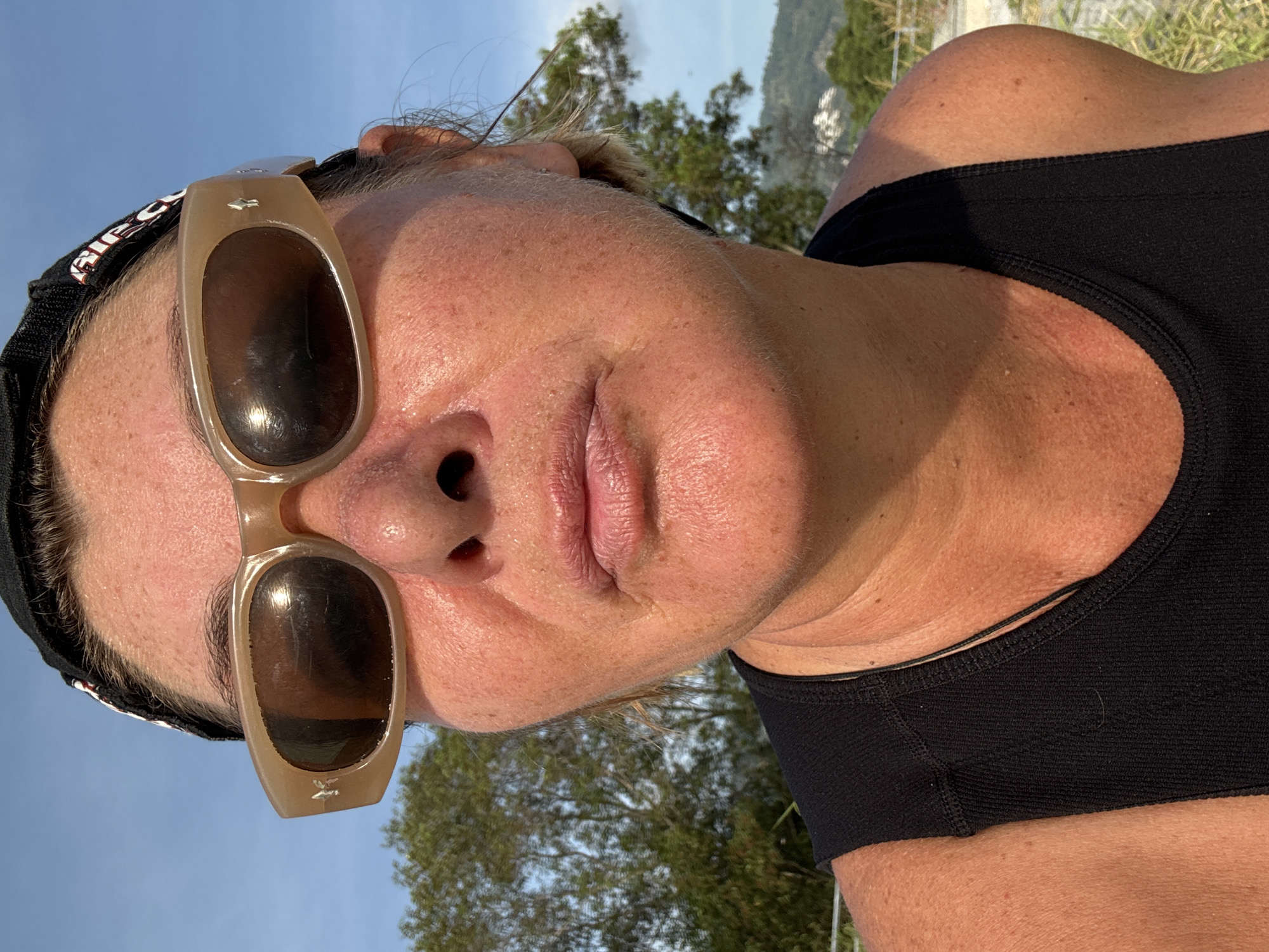 A close-up, angled portrait features a person, likely a woman, outdoors under bright sunlight. The subject is seen from the chest up, with their head tilted, filling much of the frame. They wear light brown-framed sunglasses with dark lenses, pushed up slightly on their forehead, reflecting a clear blue sky and green trees. A black headband or cap with partially visible text is worn, and the person also wears a black athletic top or sports bra.
The strong sunlight highlights the texture of their skin, showing pores and some sun-induced redness, suggesting a warm and clear day. A small star-shaped emblem is visible on the frame above the left lens of the sunglasses. In the background, beyond the subject, and especially in the reflections on the sunglasses, are lush green trees against a vibrant blue sky, indicating a natural, possibly hilly, environment. The setting is consistent with บ้านบางรักษ์, Thailand. The visible text on the black headband appears to be "RIEG". The overall impression is one of relaxation in a sunny, outdoor location.