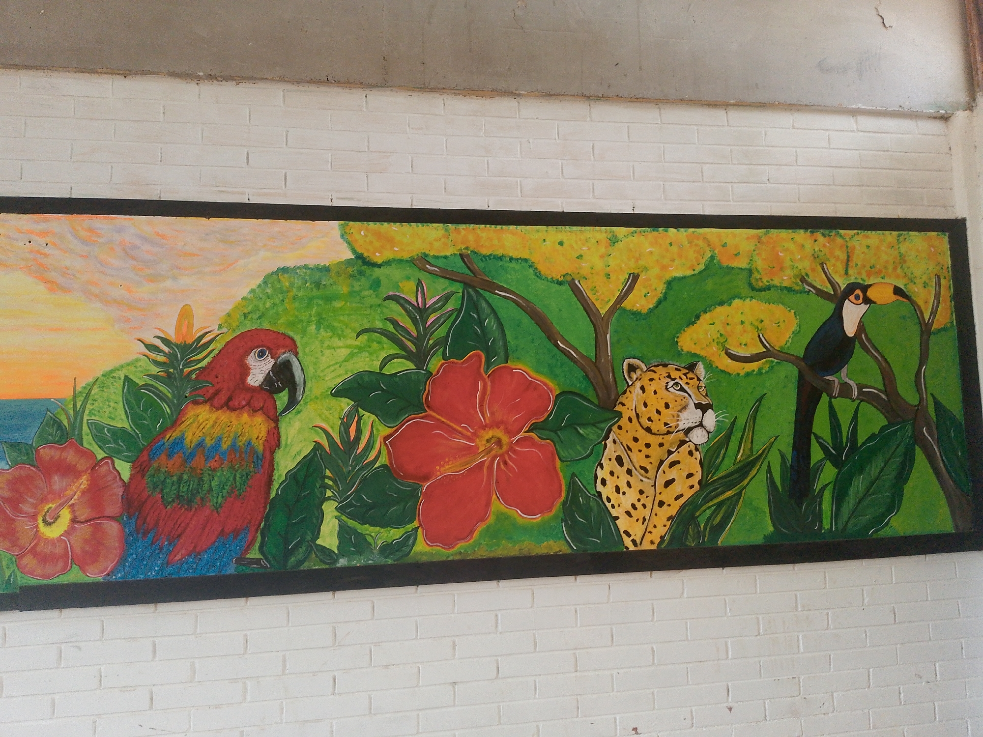 The image shows a rectangular mural with a black frame, painted on a white brick wall. The mural depicts a tropical scene with a parrot on the left, a jaguar in the center, and a toucan on the right. There are tropical flowers and plants around the animals. The background features a sunset on the left and a green and yellow forest on the right. The mural is located in Juan Griego, Venezuela.