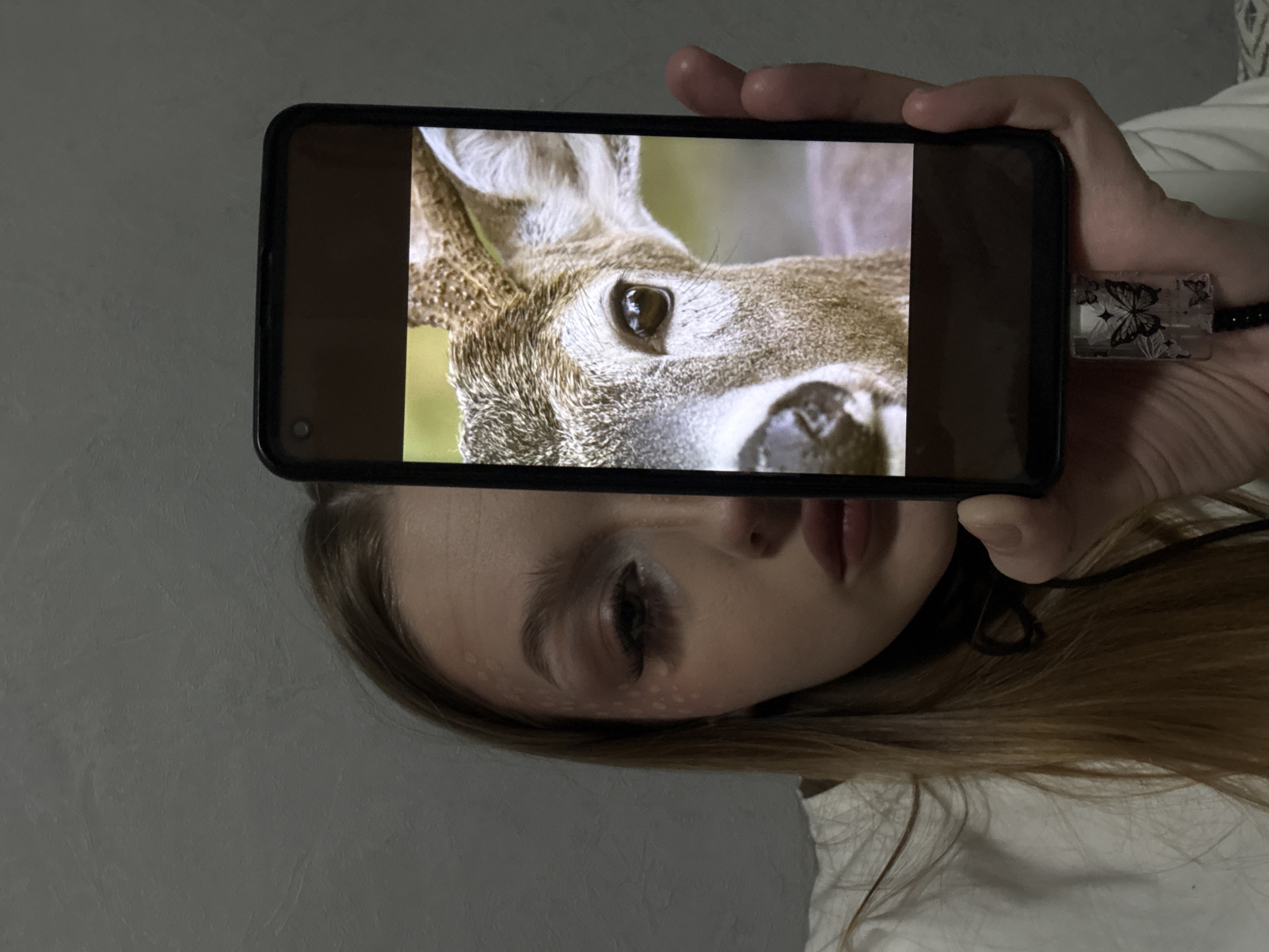 A person is holding a smartphone up to their face, displaying a close-up image of a deer's face on the screen. The deer's eye and snout are prominently visible on the phone's screen. The person holding the phone is partially visible below it, showing their eye, nose, and mouth. They have light skin and long hair. A USB drive with butterfly decorations is plugged into the phone. The background is a gray wall. This image does not contain sufficient information to determine its location within Chaikovsky, Russia.