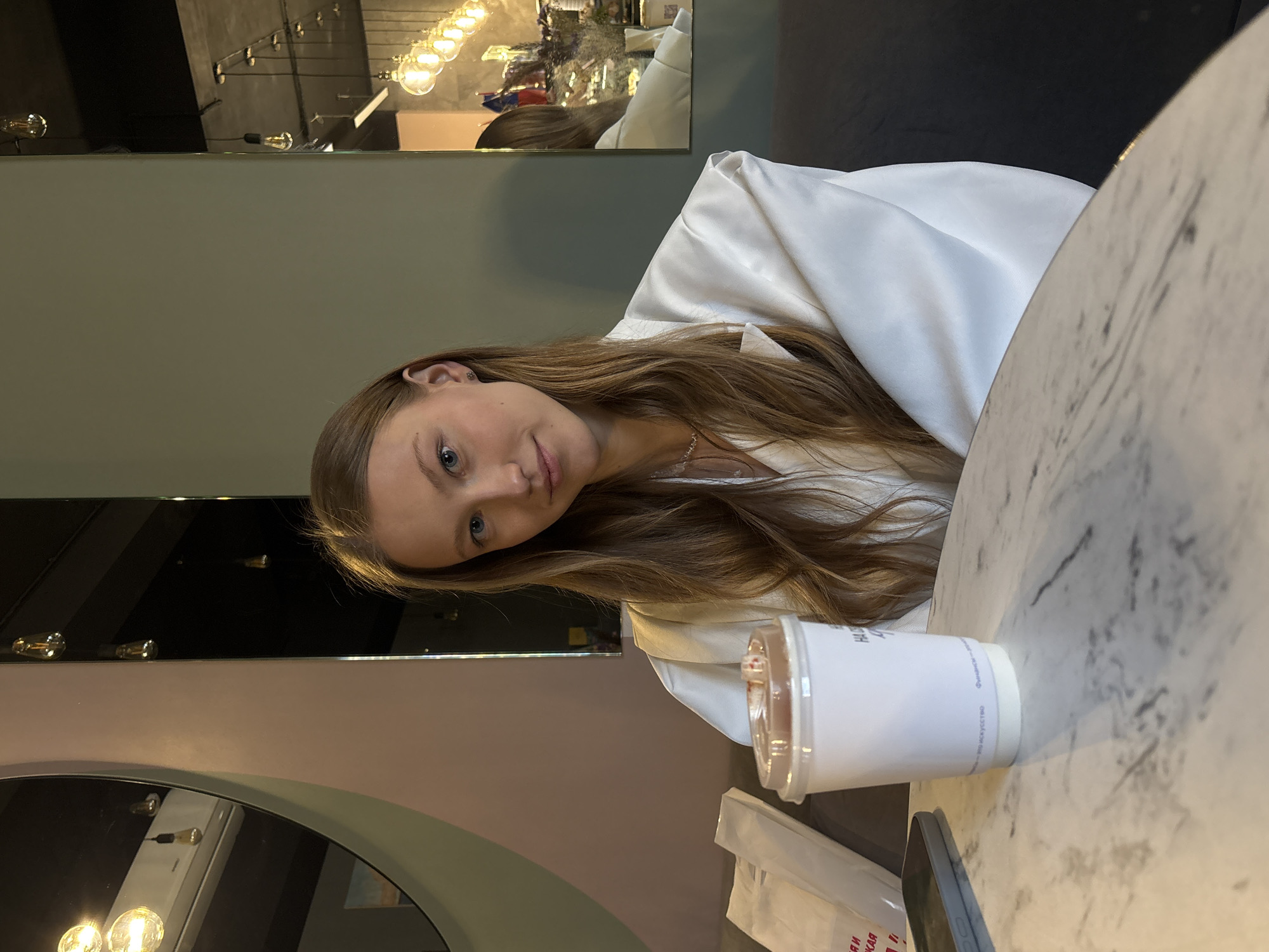 A young woman sits at a marble-topped table in what appears to be a café. She is wearing a white top. A clear plastic cup with a white sleeve, containing a beverage, sits in front of her. A mobile phone and a folded plastic bag rest beside the cup. The interior has a dark ceiling with decorative lighting and a mirror. A round mirror with lighting below is visible in the lower left corner. The image was taken indoors in Perm, Russia.