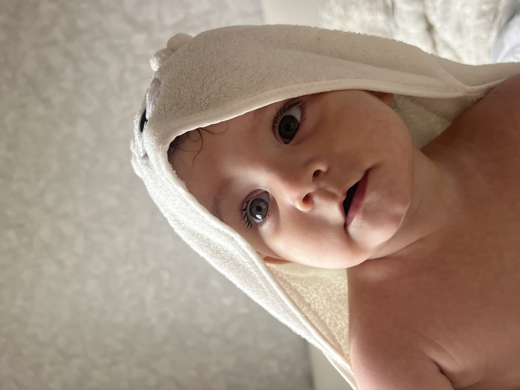 A close-up, tilted shot shows a baby wearing a white hooded towel. The baby's face is the primary subject, featuring large, expressive eyes and a slightly open mouth. The towel is positioned around the baby's head like a hood, obscuring most of the baby's body. The background is a blurred, light-colored surface. The image does not provide any geographical context to suggest Elena, Bulgaria. The subject is the baby and the object is the towel, the action is the baby looking at the camera and the environment is a light-colored background.