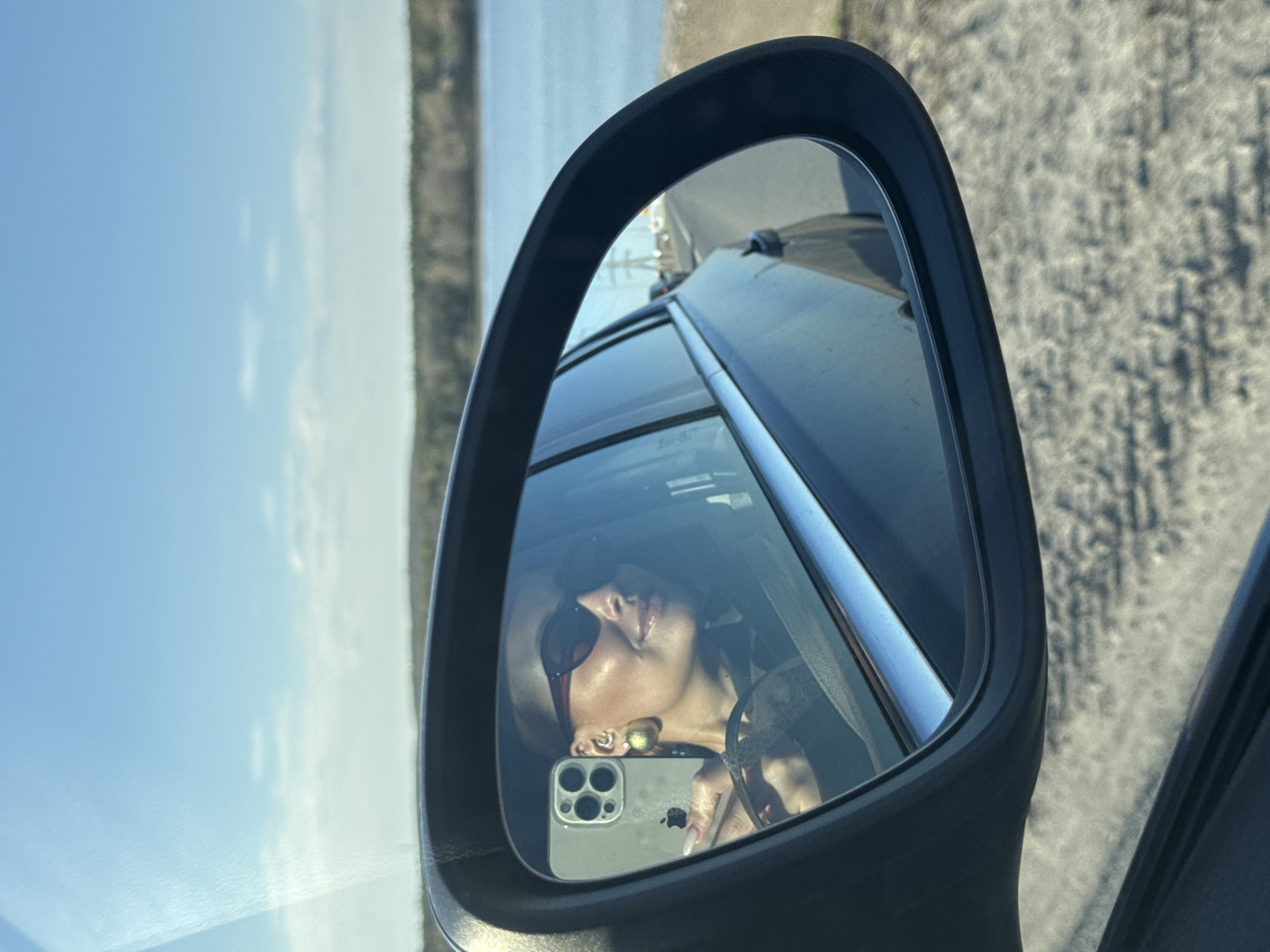 A selfie of a woman is visible in the side mirror of a car. The reflection also shows the driver, the car's interior, and the outside environment, which includes a body of water, land, and a road. Based on the context provided, this location is assumed to be Chaikovsky, Russia, although this cannot be definitively confirmed from the image alone.