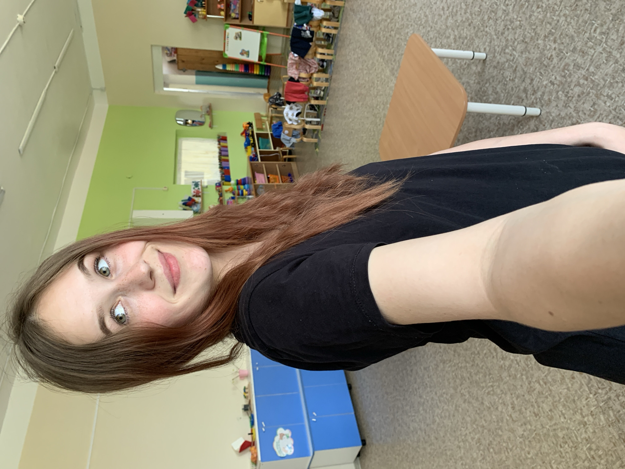 A young woman with light skin, brown hair, and freckles is taking a selfie indoors. She is wearing a black t-shirt. Behind her is a classroom setting. There are blue lockers near the bottom of the frame and a table with light brown surface on the right. The wall behind the woman is painted light green, and there is a colorful arrangement of toys and school supplies on shelves in the background. The location appears to be in the city of Фоки, Russia based on visual characteristics.