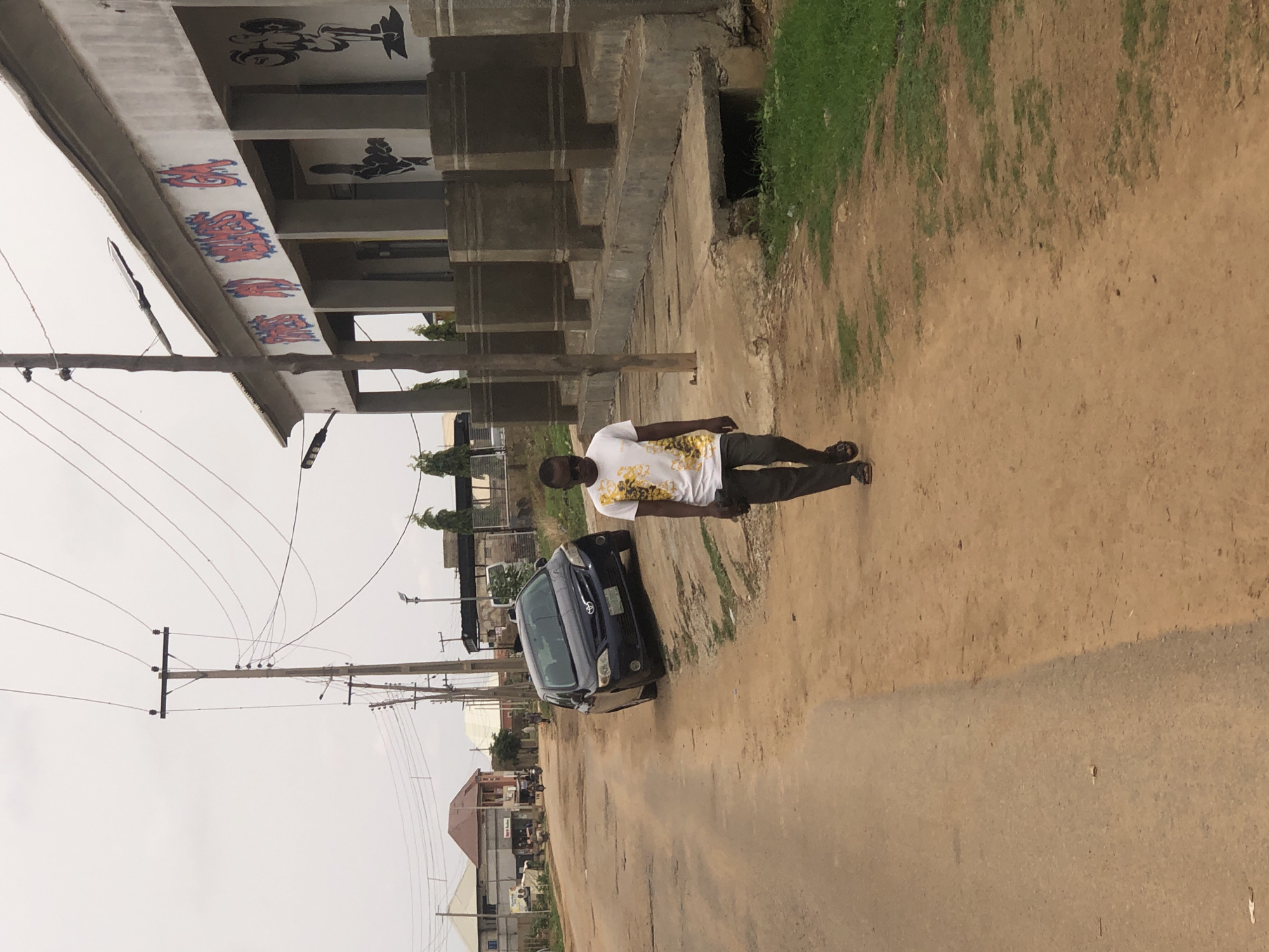 The image shows a street scene in what appears to be a residential or commercial area of Hanwa, Nigeria. A man wearing a white shirt with a gold design and dark trousers is walking along a dirt road or unpaved shoulder of the road. A dark-colored car, possibly a Toyota Corolla, is parked on the side of the road, oriented in the same direction as the man's movement. Overhead power lines stretch across the sky. In the background are buildings, some with corrugated roofing, and what appears to be a partially constructed structure with exposed concrete. The ground is mostly dirt and some patches of grass are visible. The sky is overcast. The image suggests a day time setting.