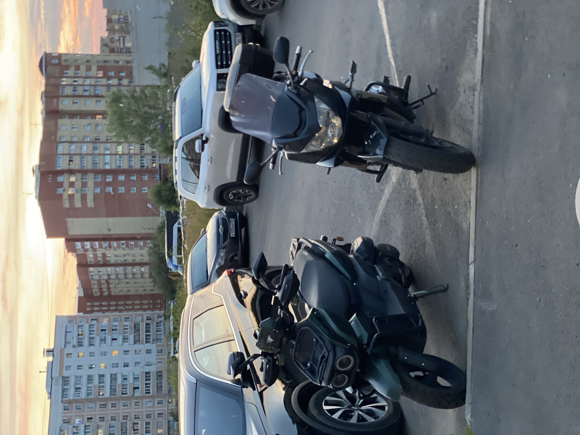 An outdoor, low-angle, eye-level shot captures a parking lot with several vehicles and apartment buildings in the background. Two motorcycles are parked prominently in the foreground. The motorcycle on the left is a dark green scooter with a black seat, black wheels, and a black front rack. It has a kickstand down. The motorcycle on the right is a black sportbike with a dark tinted windshield, black handlebars, black wheels, and a black storage box mounted on the rear. It also has its kickstand down.
Behind the motorcycles, a dark-colored SUV is parked to the left, and a lighter-colored SUV is parked to the right. A dark sedan is visible between the dark SUV and the lighter SUV. In the background, multi-story apartment buildings are visible. The sky above has a mix of pink, orange, and yellow hues, suggesting either sunrise or sunset. The ground is paved asphalt, with white parking lines visible. The scene appears to be in a residential area. The specific location in Cherepovets, Russia, cannot be determined from the visual information alone.
