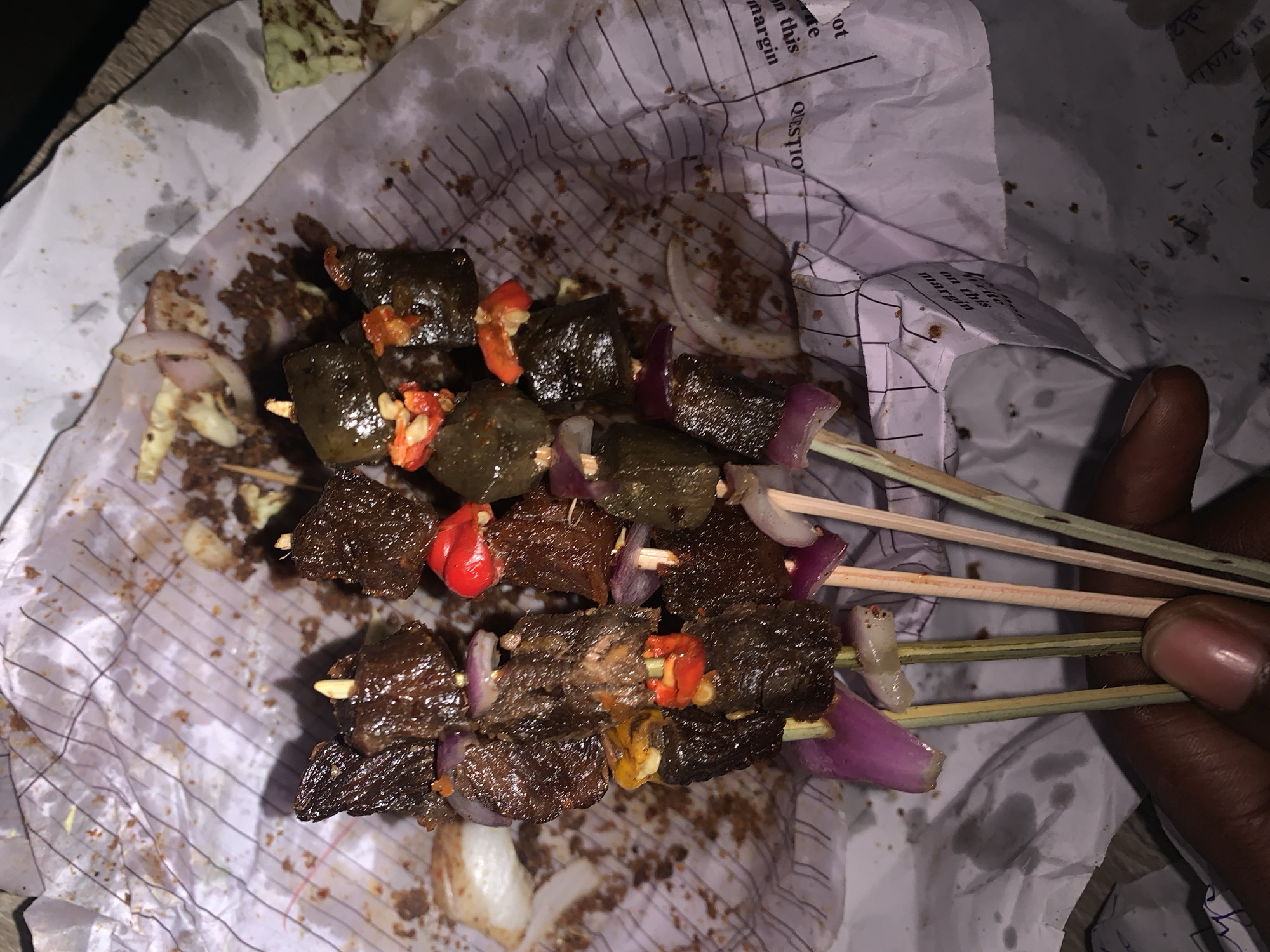 The image shows three skewers of suya (spiced grilled meat) being held. The suya consists of small pieces of dark, grilled meat interspersed with slices of red onion and pieces of red and yellow pepper. The skewers are made of thin wooden sticks. They are being held over crumpled paper with a lined pattern and text, likely used as a serving sheet. There is an oily residue and small crumbs scattered around the suya. The background includes what appears to be a piece of vegetable or salad. A hand is holding the skewers, and based on the prompt location, the setting is inferred to be near the New Stadium in Nigeria.