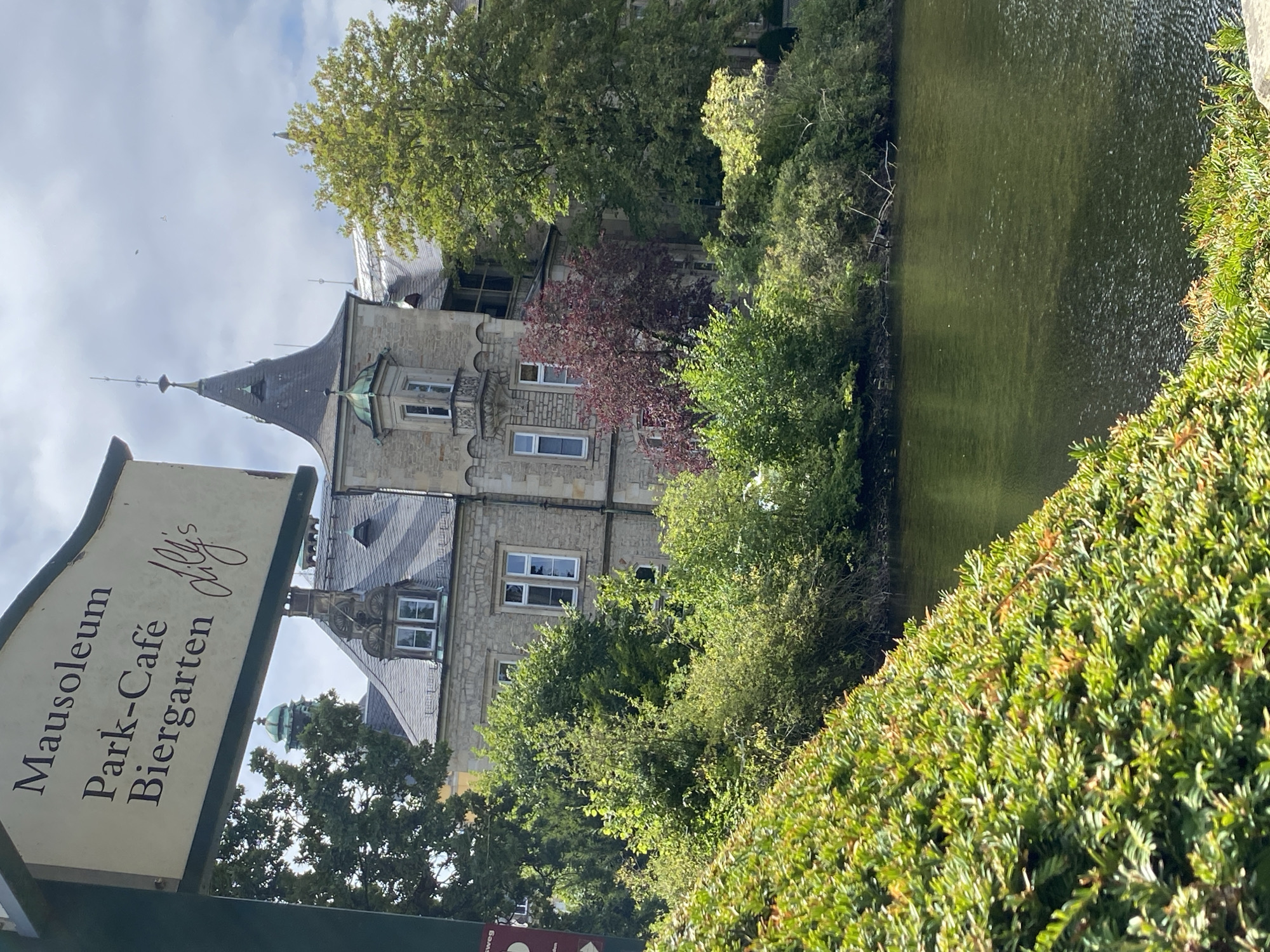The image depicts a sign in German that reads "Mausoleum Park-Café Biergarten" and a stylized signature that appears to be "dilly's." The sign is positioned in front of a lake or pond, with lush green foliage in the foreground. In the background, a large stone building with a distinctive turret and pitched roof is visible, surrounded by trees. This building is known as the Mausoleum of the Schaumburg-Lippe family, located in the Bückeburg Palace Park in Bückeburg, Germany. The sky is partly cloudy.