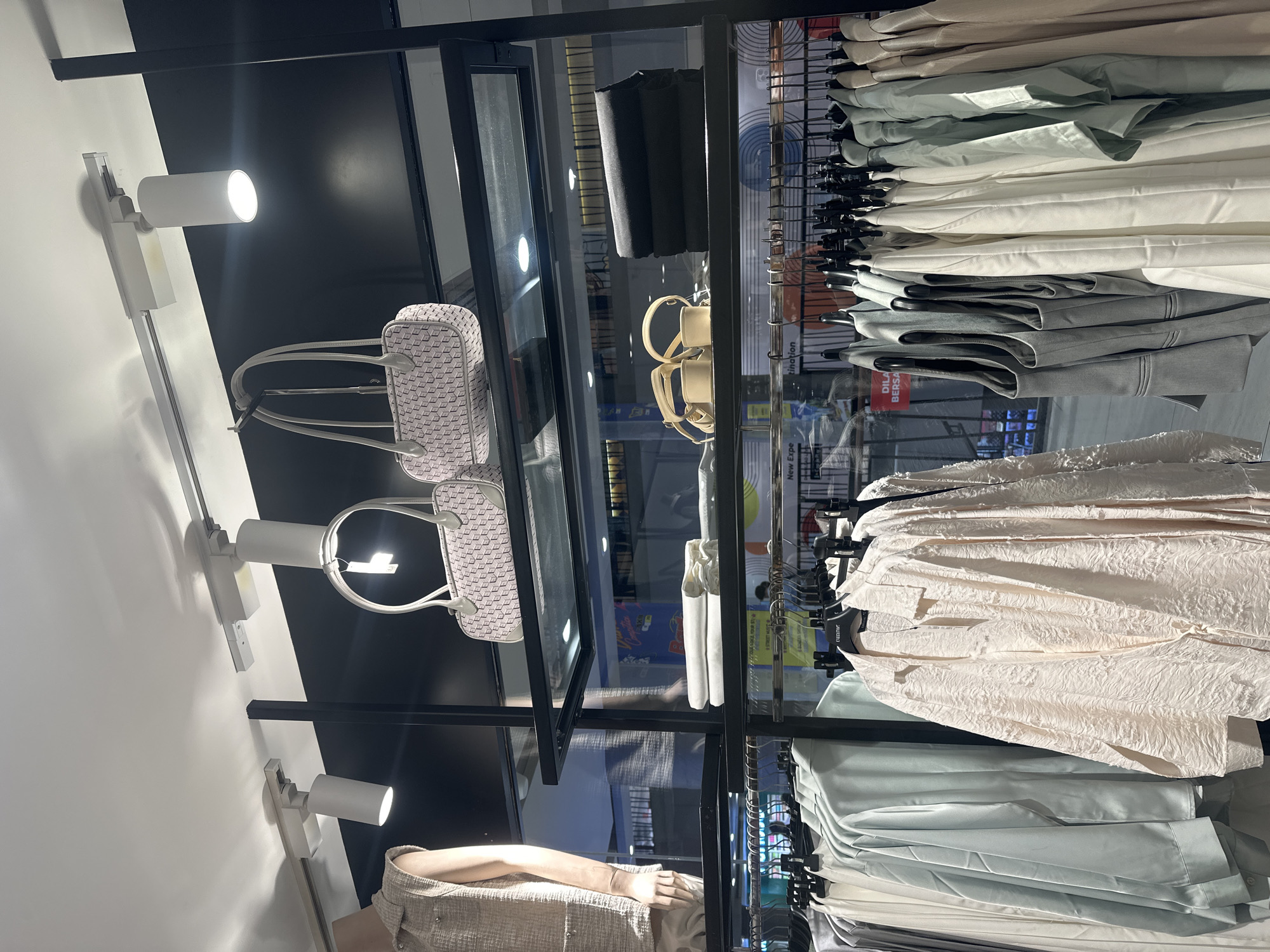 This image captures a display of women's clothing and accessories within a retail store. The scene is well-lit, with track lighting illuminating the merchandise. On a shelf above, two light-colored handbags with a repeating pattern are displayed, alongside a pair of beige sandals. Below this, several shelves are filled with clothing items. On the lowest visible rack, a variety of garments are hung, including light-colored shirts and dresses, some in pastel shades of blue and green, and others in a pale pink, textured fabric. One of the shirts, a pale pink textured garment, is prominently featured in the foreground. Other items include tailored vests in grey and more light-colored garments hanging to the right. The background of the store is visible, showing more shelving and lighting, suggesting a retail environment. A sign in red and white is partially visible in the background, which, in the context of Denpasar, Indonesia, might indicate store promotions or directional information. The overall impression is of a fashion boutique showcasing contemporary women's wear.