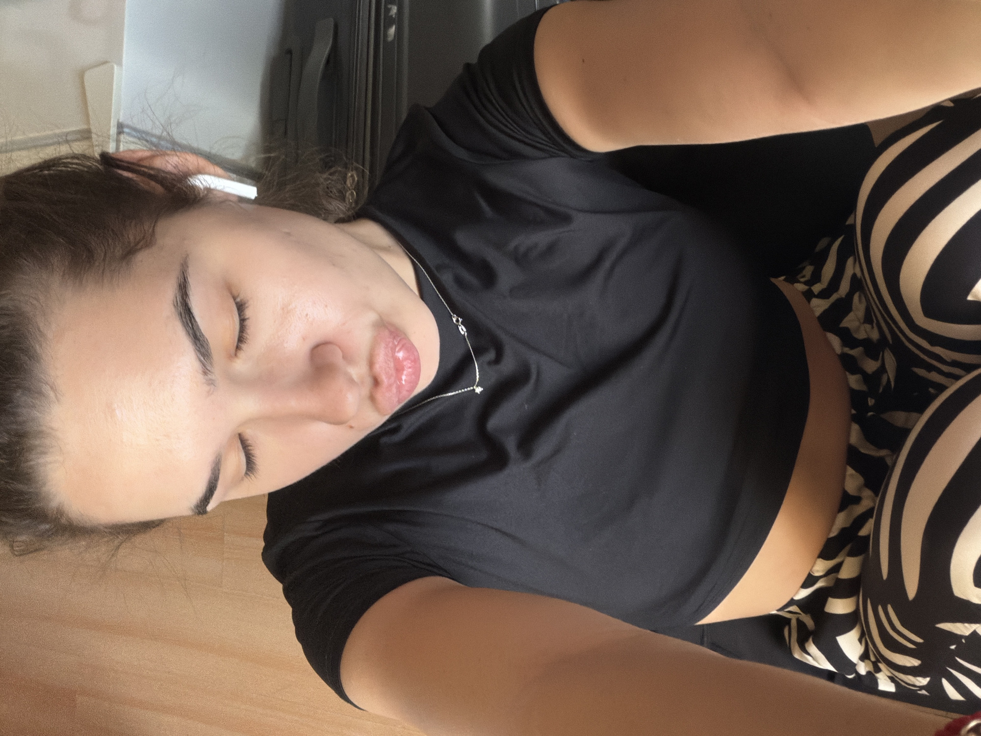A woman is pictured indoors from a close, high-angle perspective. She has light skin and dark hair pulled back, and her eyes are closed. She wears a black short-sleeved shirt and zebra-striped bottoms. A thin silver necklace adorns her neck. Background elements include a wooden floor, a suitcase, and part of a white wall. There is no information to indicate that this image was taken in the city of Sofia, Bulgaria.