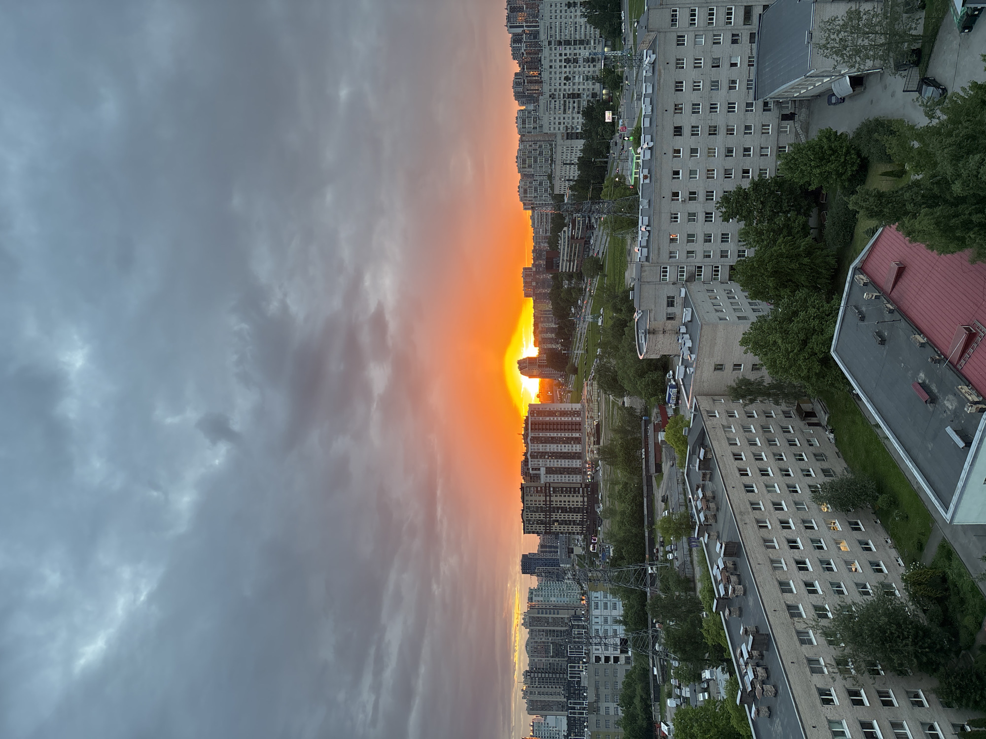 A vertically-oriented shot depicts a cityscape where a vibrant orange sky bisects the view. On the right, the urban landscape features several buildings of varying heights and architectural styles, mixed with green trees, and glimpses of roads with vehicles. The buildings have numerous windows, some showing interior lights. On the left, the sky is filled with heavy, gray clouds that contrast sharply with the bright orange of the center. The overall impression is a sunset or sunrise scene amidst a dense urban environment. The location is Unknown, Unknown.
