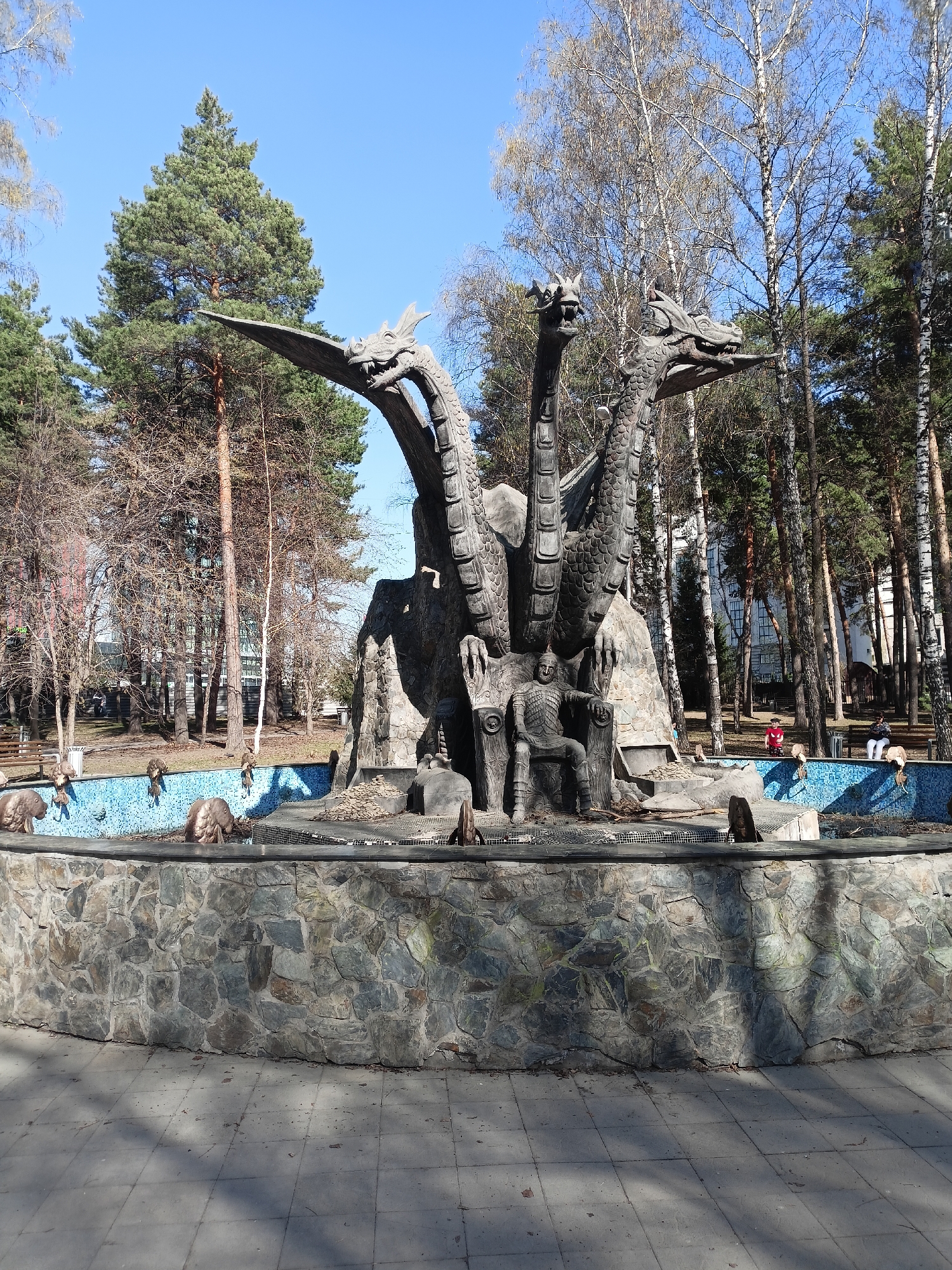 The image depicts a stone sculpture of a three-headed dragon surrounding a seated knight on a throne. The sculpture is situated in the center of a round fountain with blue mosaic tiles. A stone wall surrounds the fountain's perimeter. The area around the fountain is paved. In the background, there are deciduous and evergreen trees, and a building is partially visible. Two people are sitting on a bench in the background. The location is in Novosibirsk, Russia.