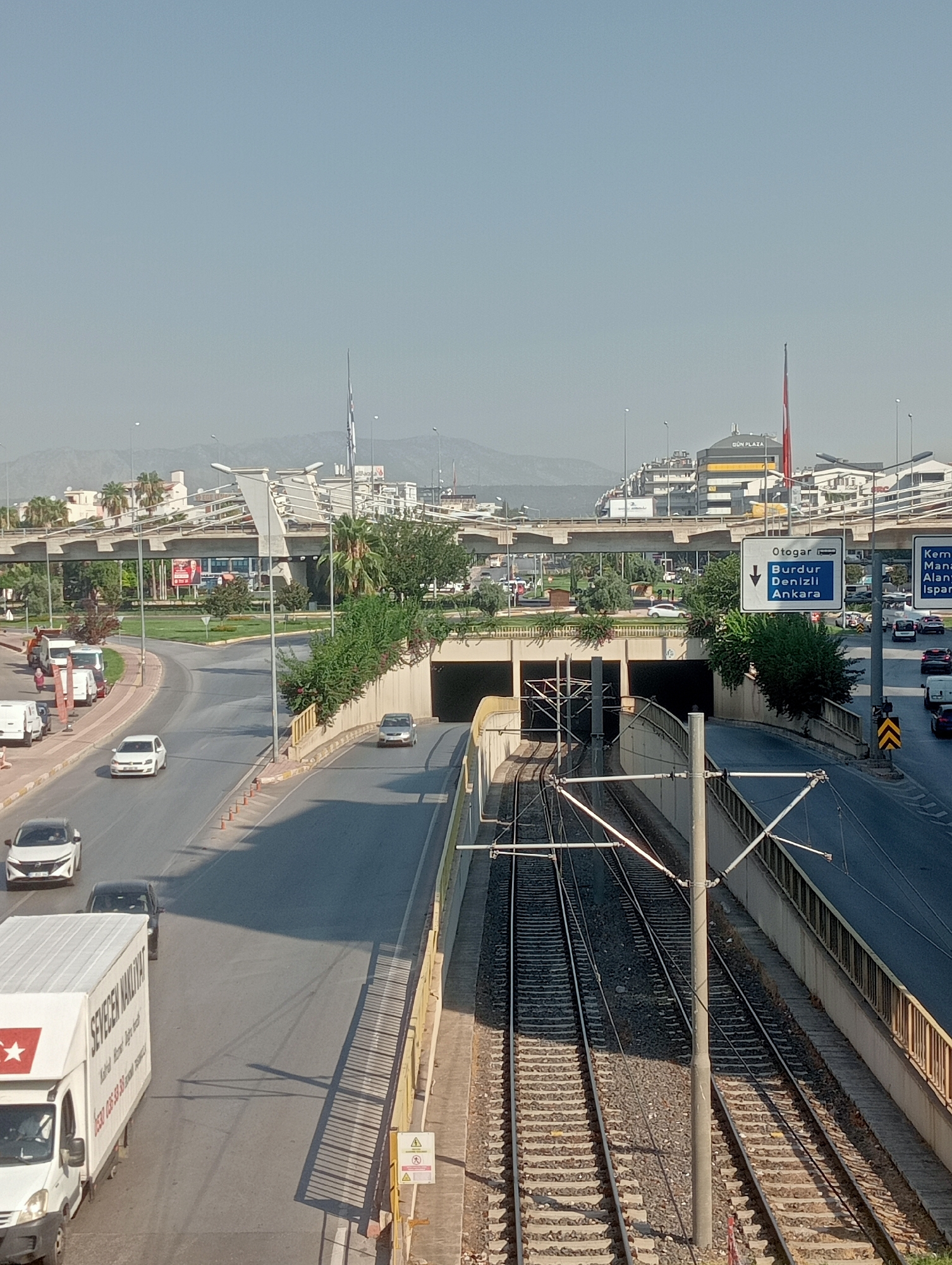 The image shows an overpass in Antalya, Turkey, likely on a highway or major road. The overpass is a complex structure with multiple levels and supports, characteristic of modern urban infrastructure. Below the overpass, a tunnel entrance is visible, with cars driving into and out of it. To the right of the tunnel, train tracks run parallel to the road, indicating the presence of a railway line. Several vehicles, including cars and a white truck with "SEVEDEN NAKLIYAT" written on its side, are on the road. The environment is urban, with buildings and greenery visible in the background. The sky is clear and blue, suggesting a sunny day. The overall scene depicts a busy transportation hub within the city of Antalya, with a focus on the integration of road and rail infrastructure.