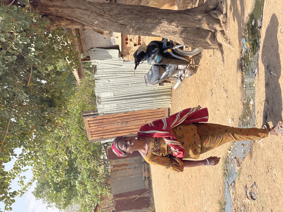 An adult female is depicted walking right-to-left on a dirt path in Chikaji, Nigeria, under daylight conditions. She wears a yellow-brown patterned top and matching trousers, a red and white patterned head covering, and a red and white striped fabric draped over her left shoulder and arm. A white bracelet is visible on her left wrist. She looks directly towards the viewer with a neutral expression. Her left foot is forward, wearing a light-colored sandal.
To her right, a dark motorcycle is parked next to a tree with a thick trunk and exposed roots. Behind the motorcycle and the woman are structures made of corrugated metal sheets, some rusted brown, forming walls or fences. A vertical wooden gate, also brown, is visible behind the woman. Further back, green foliage from trees and bushes is present. A small structure with a light-colored corrugated roof is visible behind the tree and motorcycle. The ground is dry dirt, with several shallow puddles containing dark liquid and scattered debris visible in the foreground. Shadows are cast on the ground from the tree and other unseen elements, indicating strong overhead sunlight.