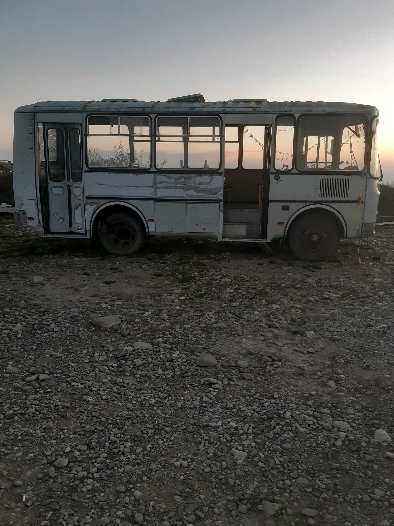 The image depicts an abandoned, light-colored bus situated on a gravel and dirt lot. The bus is weathered, with visible damage to its exterior and windows. The doors are open, revealing a dark interior. A string of small, colorful flags is draped across the roofline of the bus. The background suggests an outdoor, possibly rural setting, with a hazy sky transitioning from light to dark, indicating either dawn or dusk. The ground is covered in a mixture of small stones and dirt, with some sparse vegetation visible. The location is identified as the city of Verona, Georgia. There are no people visible in the image, and no specific actions or interactions are depicted.