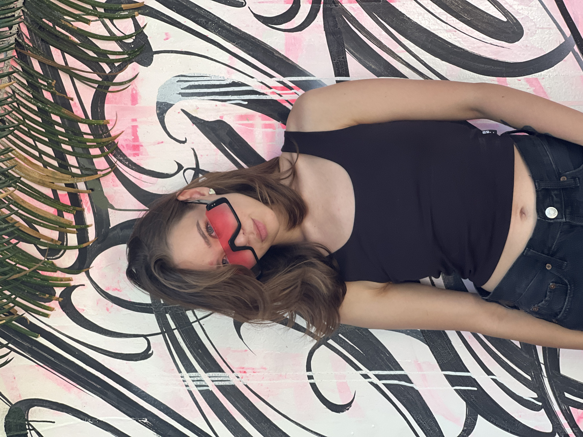 A young woman is depicted lying diagonally across the frame, her head positioned towards the upper left and her body extending towards the lower right. She gazes directly at the viewer with a neutral expression.
She has medium brown, slightly wavy hair that spreads around her head on the surface she's resting on. Her eyes are visible above a pair of distinctive, large, rectangular sunglasses with vibrant pink-tinted lenses and a black frame. Her lips are slightly parted.
She is dressed in a plain black sleeveless top, possibly a tank top or crop top, which reveals a small portion of her midriff. She also wears dark-colored, likely black or very dark blue, denim jeans, with a visible button and detail of the waistband and pocket on her right side. A small, light-colored ring is visible on her left ring finger.
The woman is lying on a surface adorned with a dynamic and abstract mural or graffiti art. The background consists predominantly of white, black, and various shades of pink. Bold, sweeping black strokes, swirls, and lines create an energetic pattern across the surface. These black elements are interspersed with and often layered over areas of bright pink, which appear as splashes, drips, and brushstrokes.
In the upper left corner of the image, dark green, thin, pointed leaves or fronds, possibly from a palm tree or similar plant, overlap the painted background. Some of these leaves show signs of drying or browning at their tips.
The lighting is bright and even, suggesting an outdoor setting during the day. The overall impression is casual and artistic, blending the subject's relaxed pose with the vibrant, urban art background.