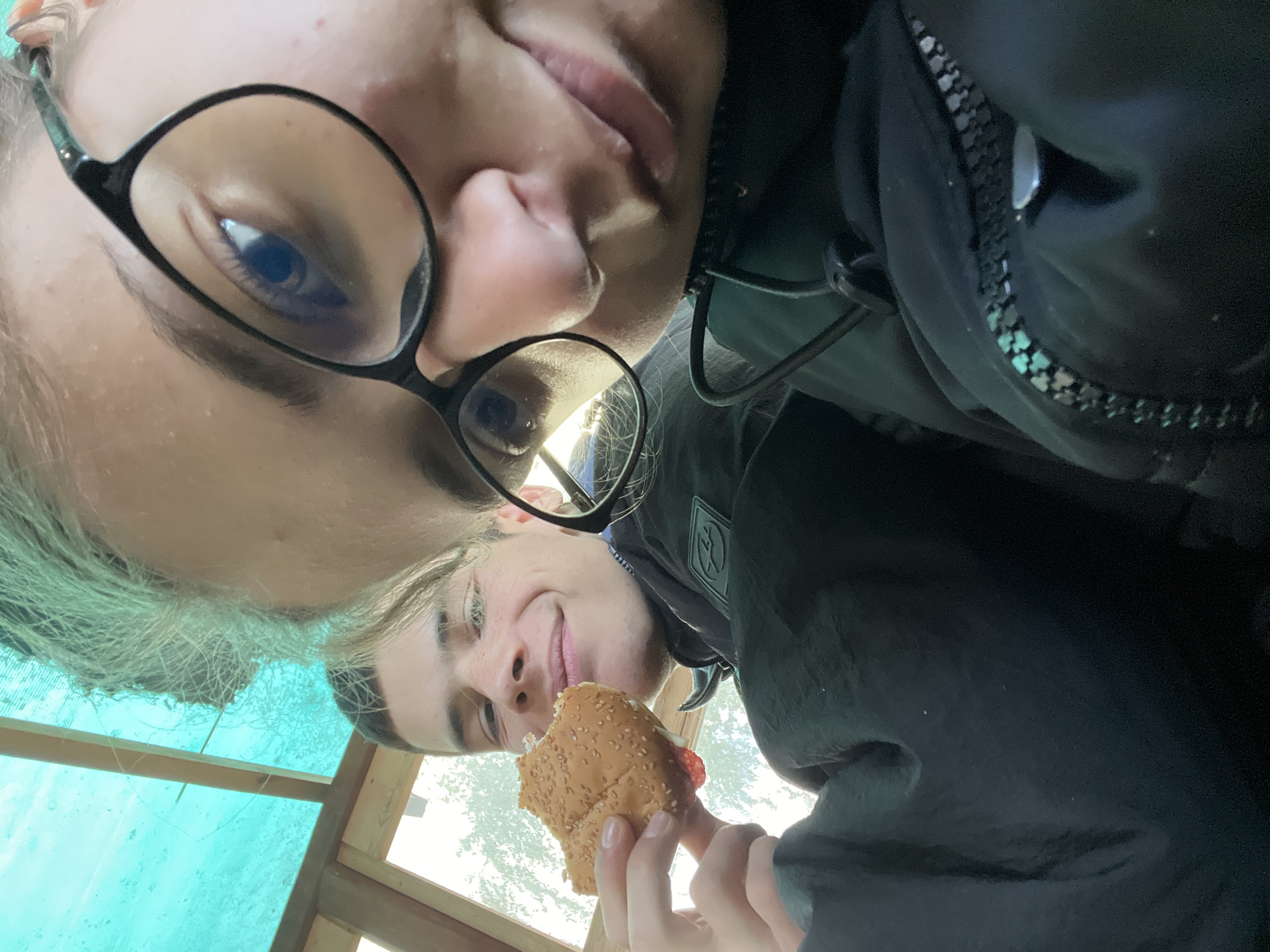 Two individuals are positioned closely together in an upside-down selfie taken at an upward angle. The individual on the left is wearing glasses and has fair hair. The individual on the right is holding a partially eaten sandwich. The background is comprised of an outdoor scene with some vegetation. The location is Karavelovo, Bulgaria.
