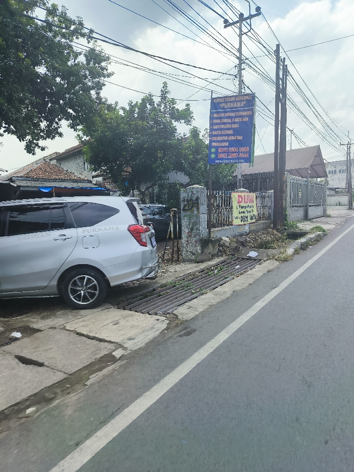 The image shows a silver car, likely a Toyota Avanza, parked on the side of a paved road in Ngamprah, Indonesia. The car is parked on the right side of the road, facing away from the viewer, with its rear end visible. There is a drain next to the car, with a metal grate. There are several power lines running overhead. There is a large blue sign on a post to the right of the car, with white text that appears to be advertising a driving school called "Purnama." There is a smaller, yellow sign underneath the blue sign. There is a building on the left side of the image, with a small, flat roof. There is a large green tree in the background. The road is empty and the weather appears to be sunny. The scene is deserted, with no people present.
