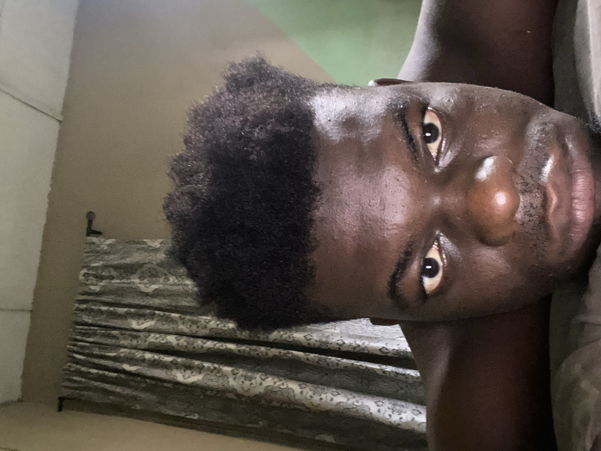 A close-up, head-and-shoulders shot of a dark-skinned person with short, dark, curly hair. The person is lying down, facing the camera. Their skin appears shiny, possibly due to moisture or oil. The background includes a bed with a gray and white patterned bedspread, a wall in a muted brown tone, and a glimpse of green in the upper right corner. The location in the city of Unknown, Unknown is not discernible from the image.