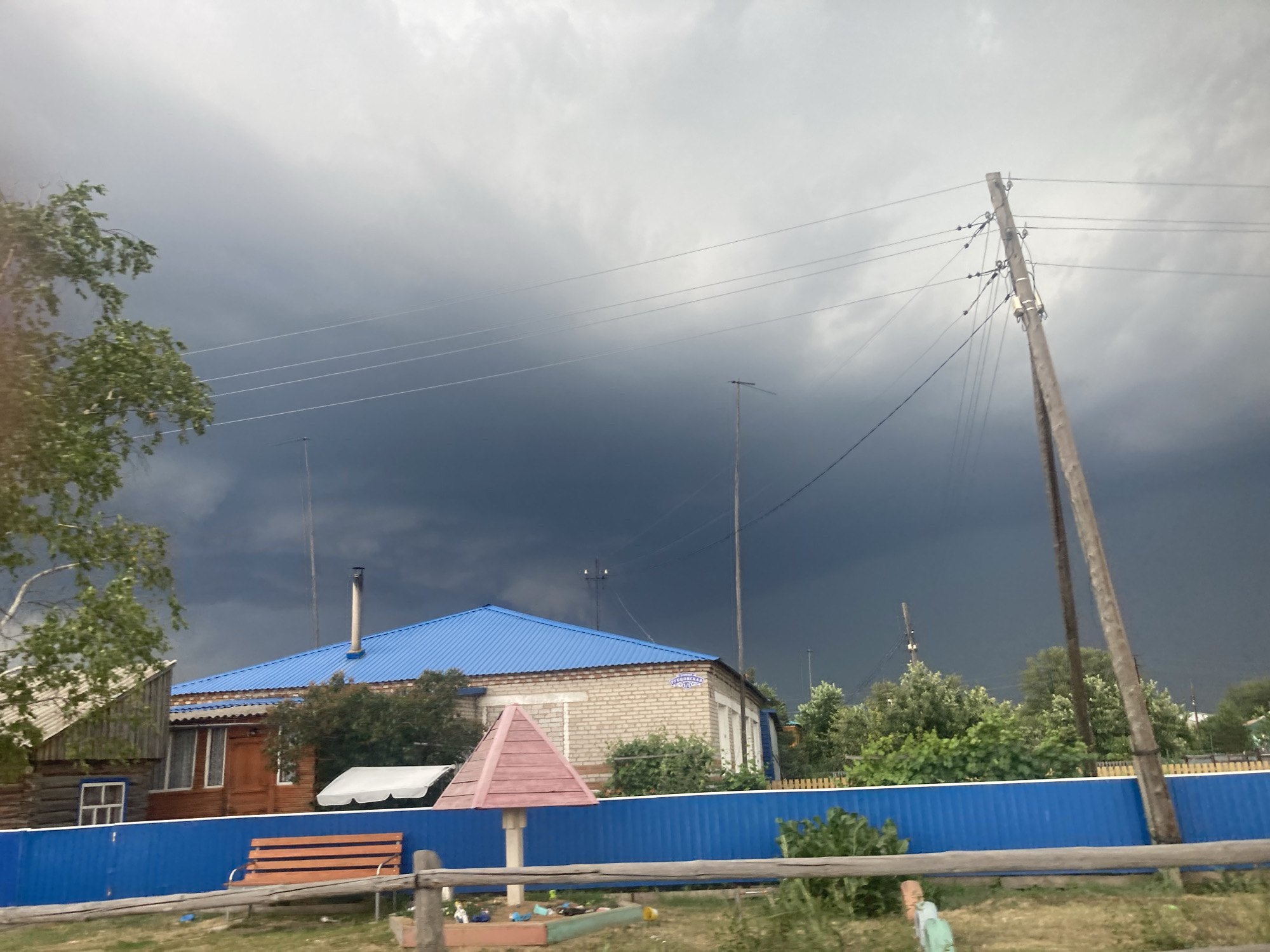 The image depicts a rural scene in Kormikha, Russia, under a dramatically overcast sky. A dark, foreboding storm cloud looms overhead, casting a somber mood. In the foreground, a bright blue fence runs horizontally across the frame, creating a distinct visual separation. To the left of the fence, a wooden bench with orange slats is positioned on a patch of grass. Behind the fence, several structures are visible. A building with a blue metal roof and a brick facade stands prominently, with trees and foliage surrounding it. To the left of this building, a portion of a wooden house with a visible window is present. Various utility poles and wires are scattered throughout the scene, with one particularly tall wooden pole dominating the right side of the frame. A small, pink, pyramid-shaped structure, possibly a child's playhouse or decorative element, is situated near the blue fence. The overall impression is one of an impending storm in a quiet, rural settlement. There are no people or animals visible in the image.