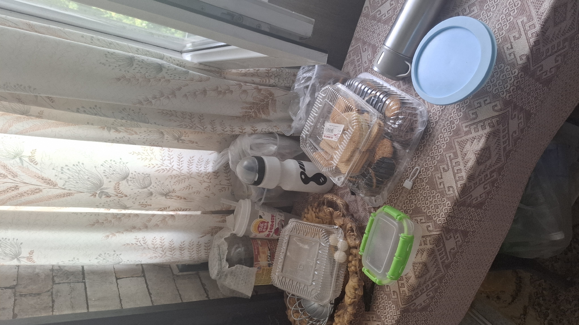 The image depicts a close-up, eye-level shot of a table covered with a patterned tablecloth. The table is laden with various food items and containers, suggesting a domestic setting. In the foreground, a green and clear plastic food container is prominent. To its right, several plastic containers hold an assortment of baked goods, including cookies and what appear to be small cakes or pastries. A light blue lid is positioned near a silver thermos.
Behind these items, to the left, is a clear plastic container with a lattice-like pattern on its side, and within it, small round white objects. Further left, a dark glass jar with a label is partially visible. A white plastic cup with a straw is situated near the jar. A white water bottle with a black lid and a black figure-eight design is also present.
The background consists of a curtain with a subtle floral or botanical pattern, drawn to the right, revealing a window through which natural light streams in. The wall to the far left is constructed of light-colored, textured bricks. The overall impression is one of casual arrangement, possibly indicating a snack or a gathering. The scene is indoors, with the light suggesting daytime. Without further information, it is impossible to definitively place this scene in Shymkent, Kazakhstan, as the image lacks any identifiable landmarks or cultural markers specific to that location.