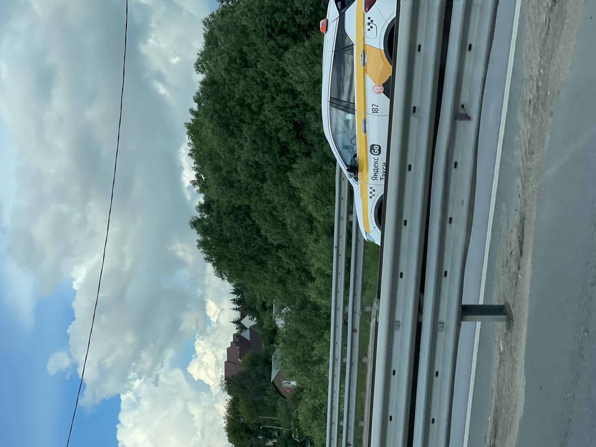 A Yandex Taxi is on the roadside barrier in Bykovka, Russia. The taxi is white with yellow striping and Yandex Taxi markings. A dense green forest is in the background, along with houses with red roofs. A cloudy sky with patches of blue is visible above. A roadside barrier runs horizontally across the lower center of the image. A power line is visible in the sky to the left of the image.