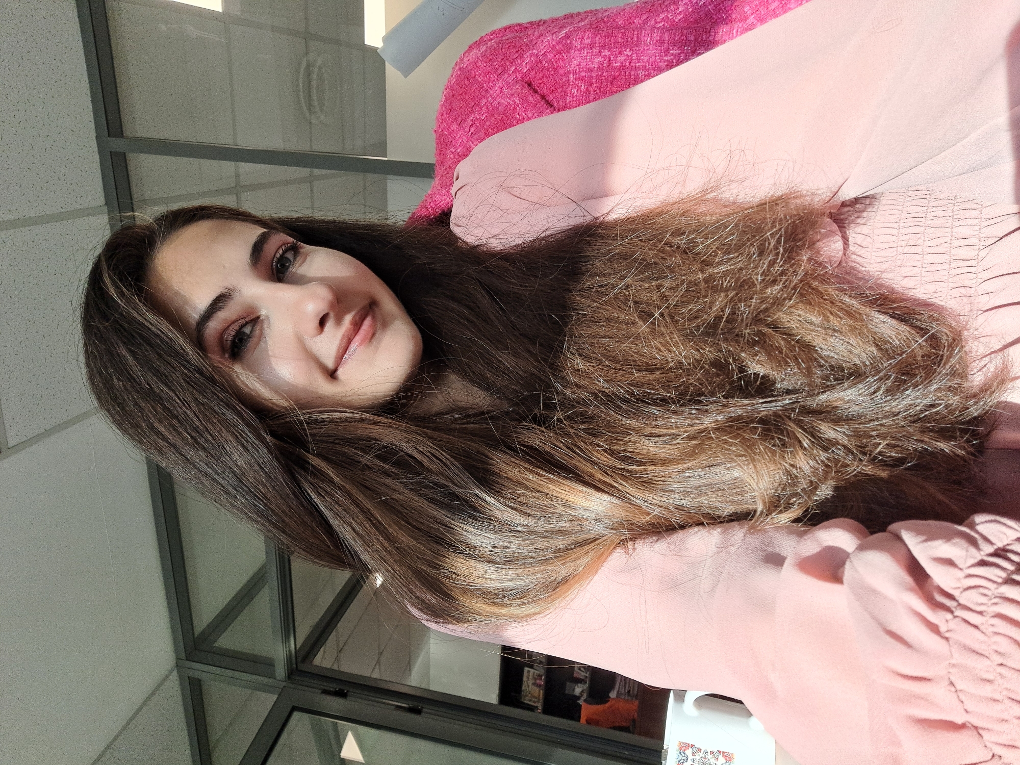 A young woman with long, dark brown hair, highlighted with lighter brown tones by sunlight, is the central subject of the image. She is looking directly at the camera with a gentle smile, her left eye, nose, and mouth visible. Strong natural light, likely from a window to her left, illuminates her face, creating bright highlights across her forehead and nose.
She is wearing a light pink top, possibly made of chiffon, featuring a shirred or smocked cuff on the visible right sleeve. Over this, she wears a vibrant, textured bright pink garment, possibly a jacket or cardigan. Her hair flows down past her shoulders, covering part of her arm and the light pink top.
The background suggests an indoor setting, likely a commercial or institutional building, characterized by large glass panels or windows with dark frames, and a ceiling with acoustic tiles. The strong sunlight indicates it is daytime. A small portion of a white mug with an indistinct colorful design is visible in the bottom right corner. No legible text is visible in the image. The overall impression is bright and pleasant, with the woman appearing calm and engaging.