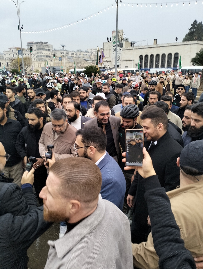 A large crowd gathers outdoors on a cloudy day. Many men are present, some wearing traditional Muslim attire, others in casual clothes. A few individuals sport bicycle helmets. Several flags with Islamic symbols are visible in the background. Buildings line the horizon, and a string of lights hangs overhead. People are holding up phones and cameras, capturing the event.