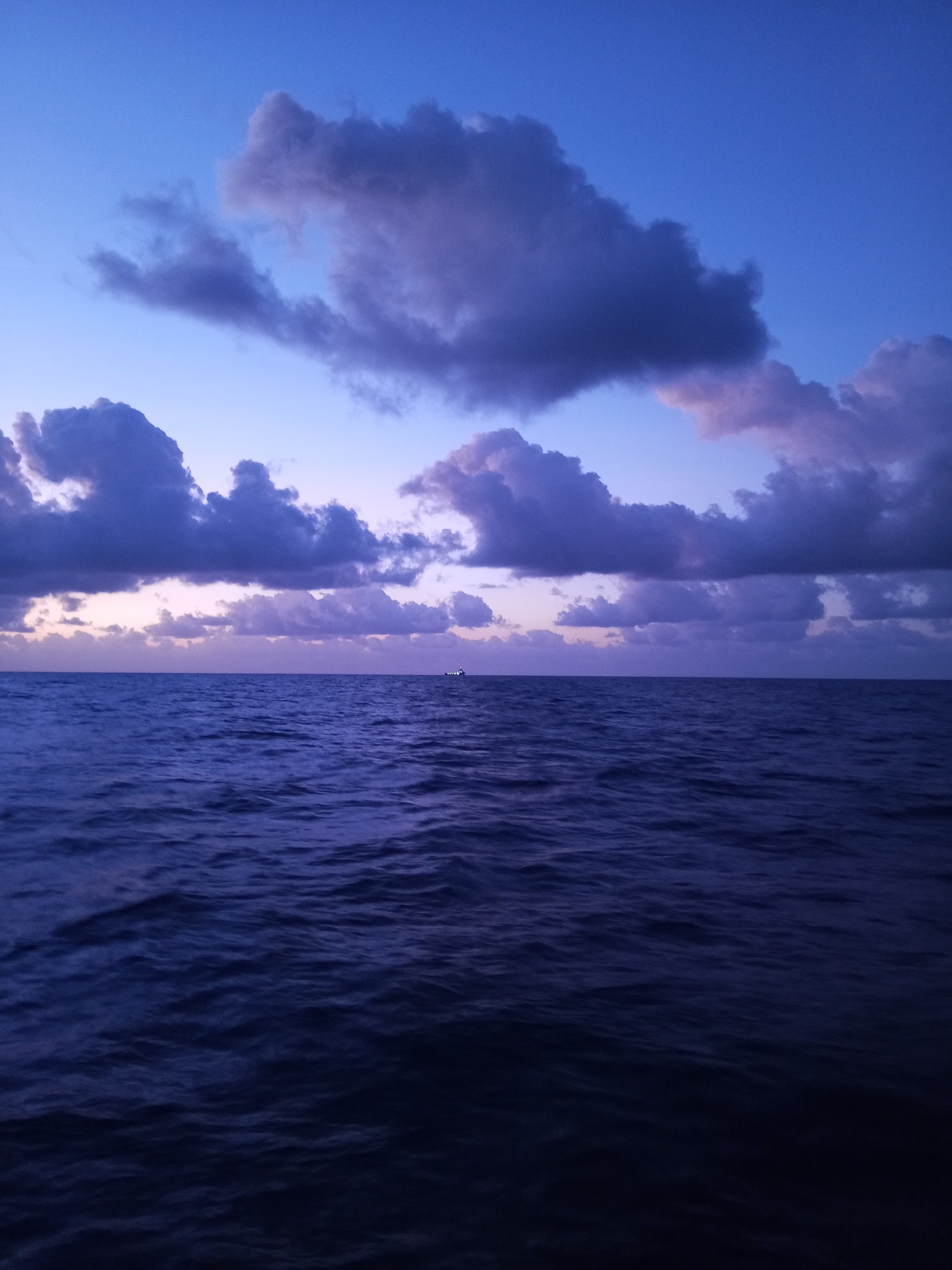 A wide shot captures the vast expanse of the ocean under a deep blue twilight sky. Wispy, dark purple clouds drift across the horizon, illuminated by the fading light. The sea is a deep, dark blue, with gentle ripples creating a textured surface. In the distance, a small ship is visible, a tiny silhouette against the dramatic sky and sea. The scene evokes a sense of calm and solitude, as if at the edge of the world.