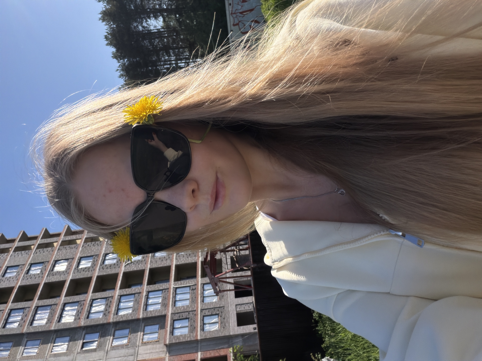 A young woman with blonde hair, wearing sunglasses and a light-colored zip-up jacket, is positioned in front of an unfinished building in Izhevsk, Russia. She has a dandelion flower tucked into her hair near her sunglasses. The building behind her has a unique architectural style with many windows and concrete exterior. Part of a treed area is visible behind and above the building. The sky is clear blue. The woman is looking at the camera and seems to be holding the camera herself.