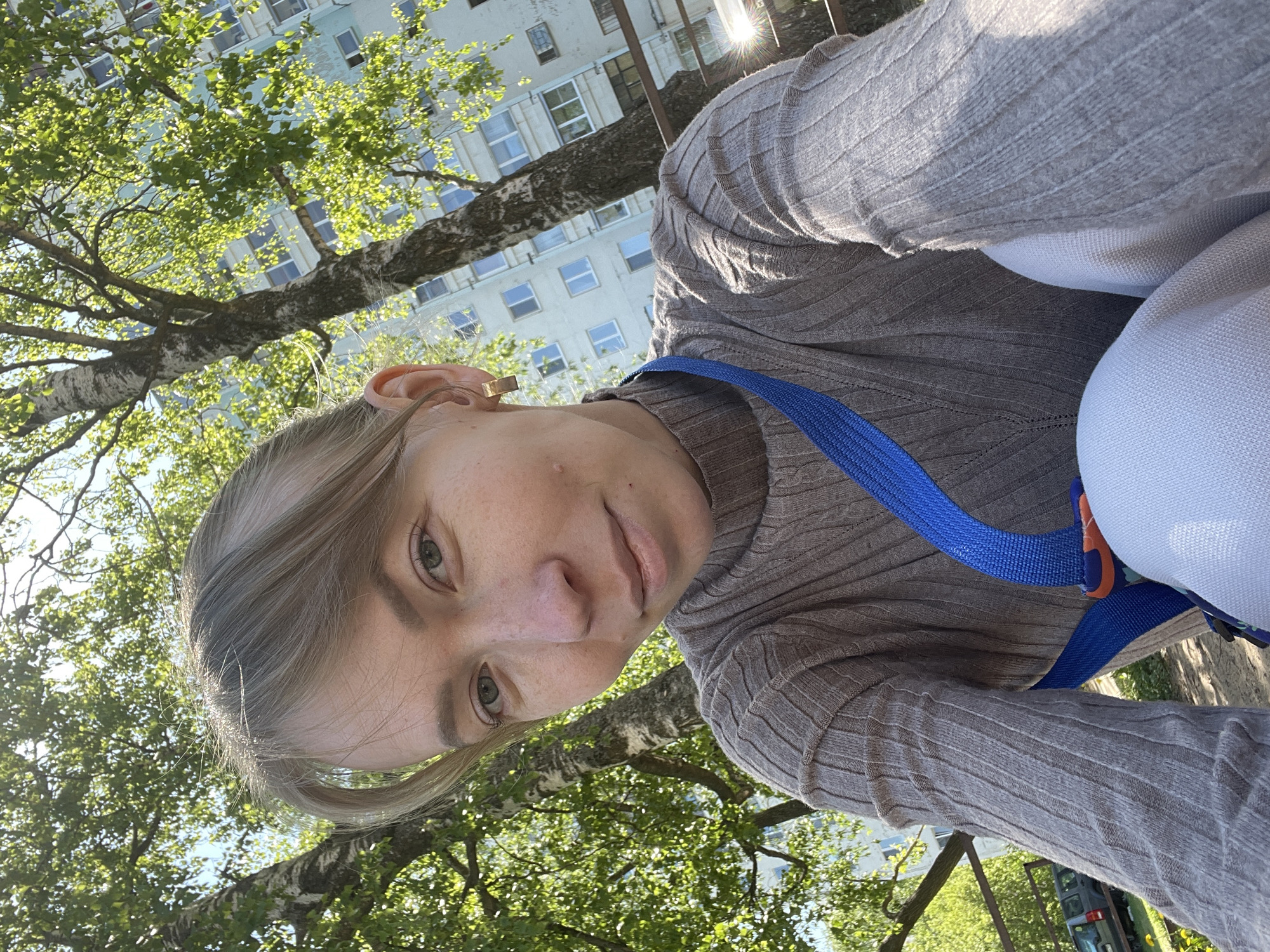The image is a selfie taken outdoors, likely in Perm, Russia, given the urban environment and foliage. A young woman with light hair and fair skin is the primary subject. She is wearing a gray ribbed sweater and a blue strap or harness, possibly for carrying something. Behind her, there are trees with green leaves and a multi-story building with numerous windows. The sun is shining, creating bright spots of light in the foliage. The perspective is angled upwards, suggesting the photo was taken from a slightly lower position.