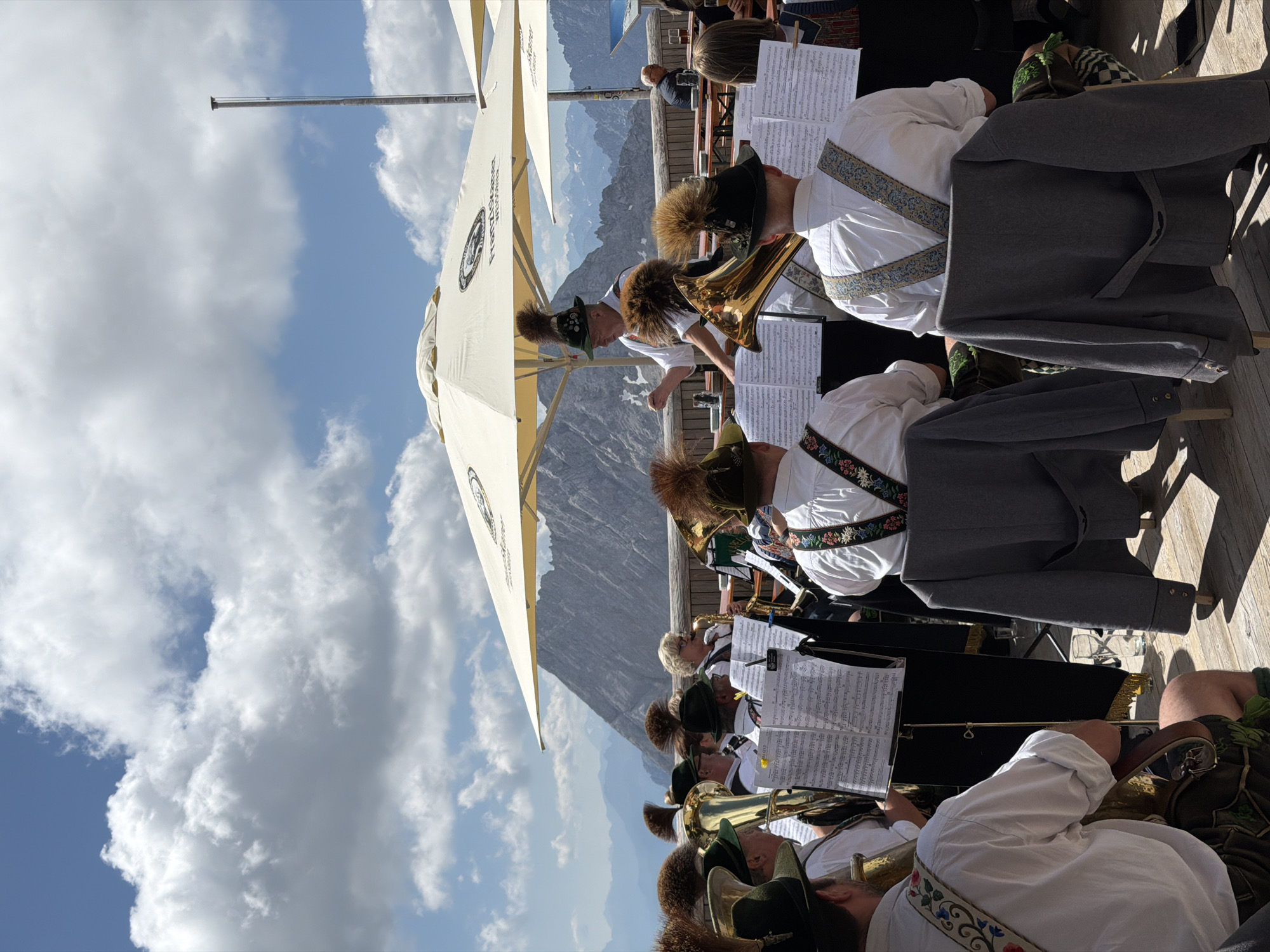 A brass band is performing outdoors on a wooden deck with a mountainous backdrop. The band members, all male, are wearing traditional Bavarian attire, including lederhosen and feathered hats. They are playing various brass instruments, such as tubas and trombones, and reading from sheet music displayed on stands. A conductor, also in traditional dress, is directing the band. In the background, there are large white umbrellas providing shade, tables and chairs, and more people seated. The sky is blue with scattered white clouds, and the mountains in the distance are rugged and snow-capped. The location is the Zugspitze, a mountain on the border between Austria and Germany, near the village of Wildermieming in Austria, suggesting a performance at a high-altitude restaurant or viewpoint.
