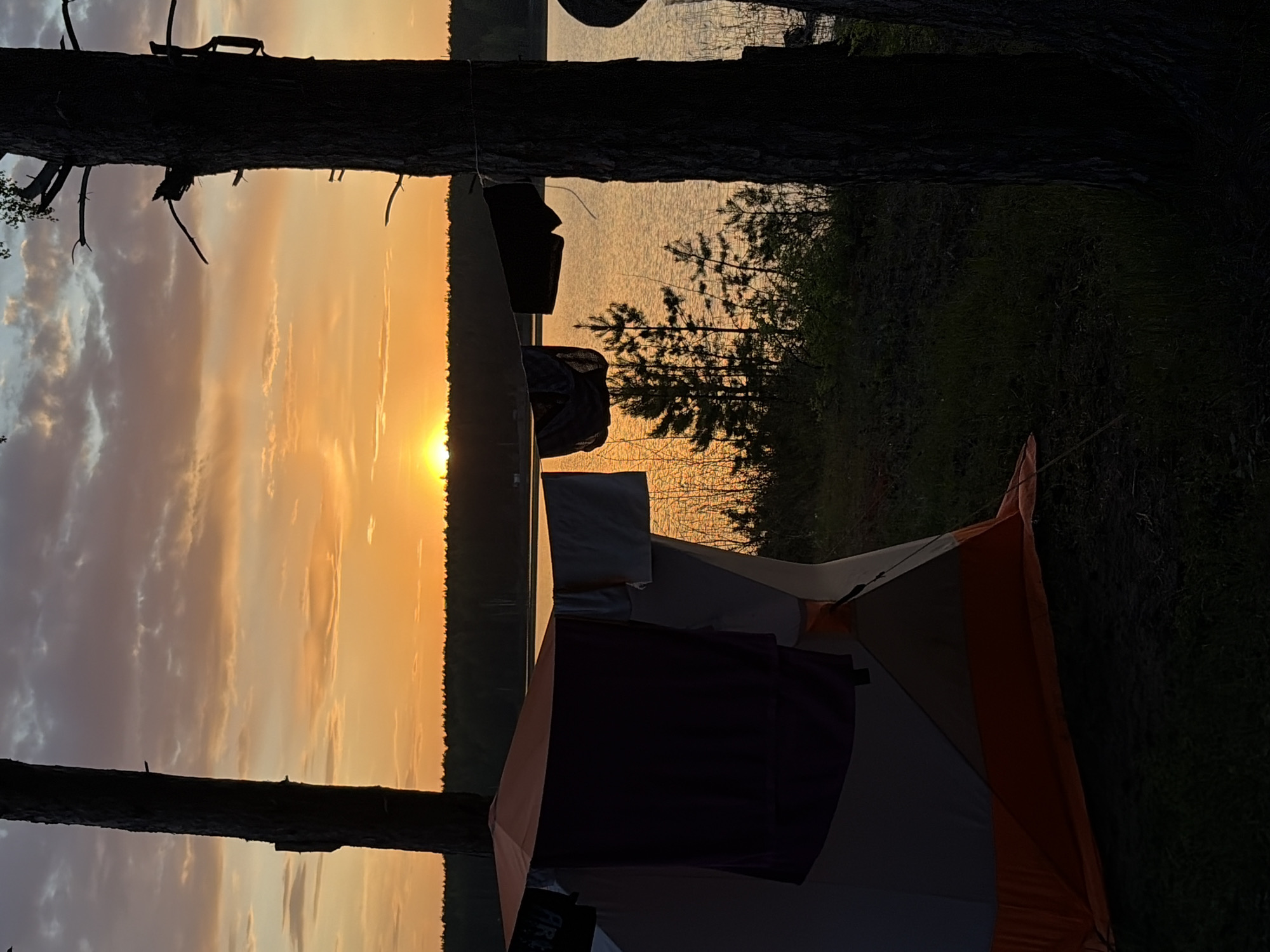 The image shows a campsite at dusk or dawn, possibly in a natural setting like Zabegaevo, Russia. A two-person tent, orange and gray, is visible in the foreground. Clothes are hanging on a line strung between two trees in the background, with a body of water beyond them and a forest edge in the far distance. The sky is a mix of golden and orange hues, indicating sunrise or sunset. The light is muted, and there's an overall feeling of tranquility.
