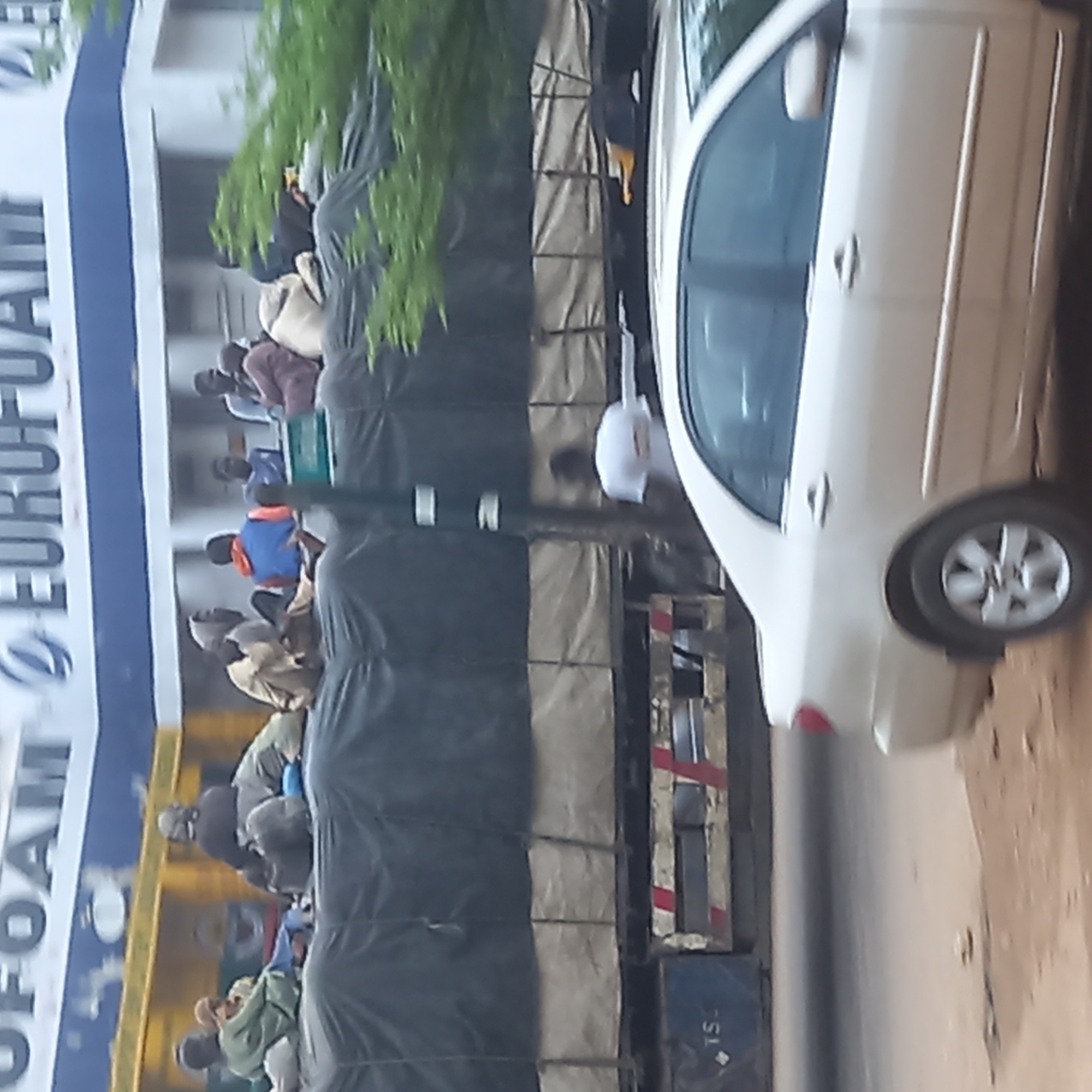 In Sardauna, Nigeria, a white car is parked beside a truck. The truck is crowded with people. The truck appears to be transporting goods. A sign for EUROFOAM is visible. The environment is urban.