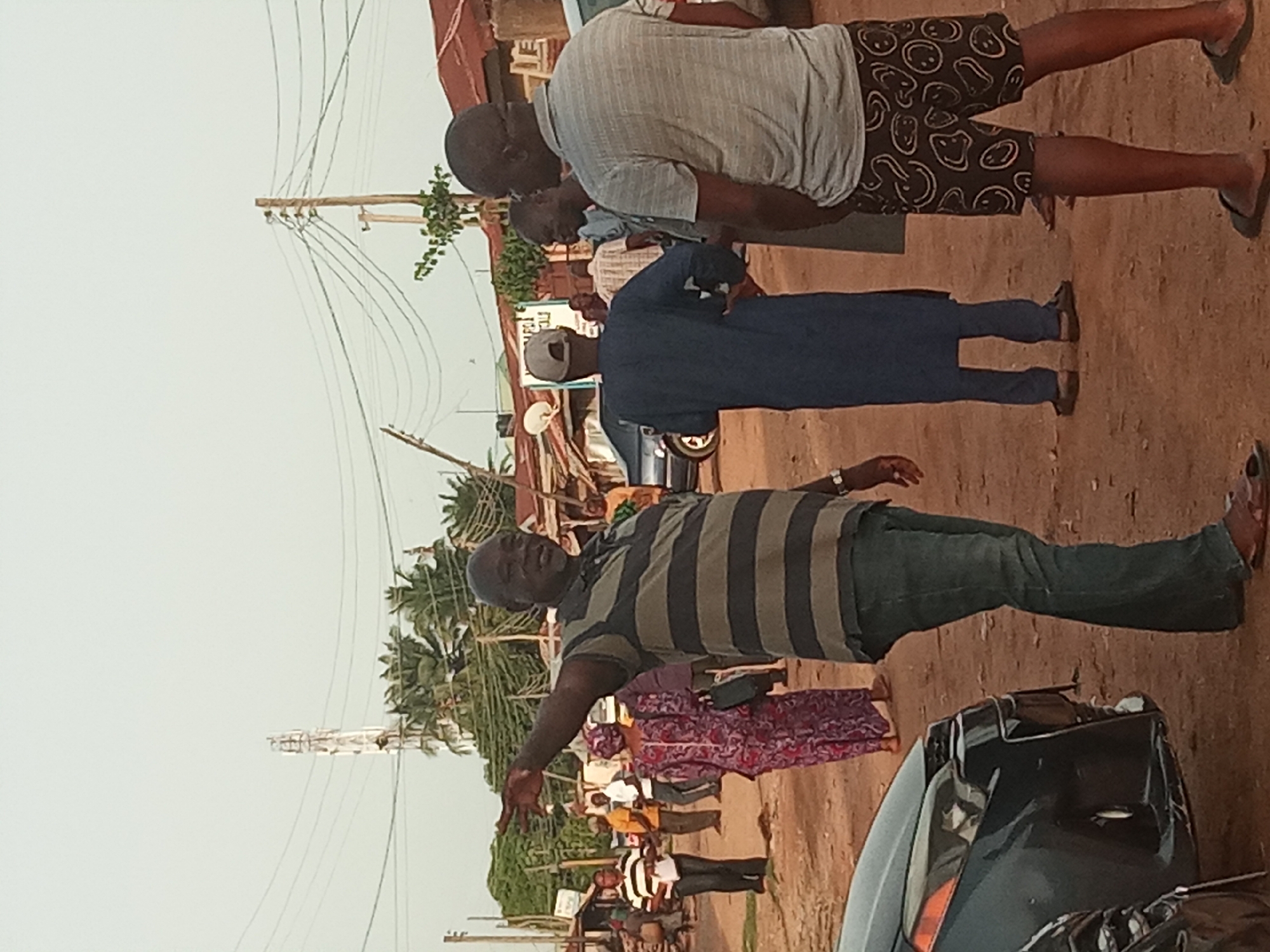 A medium shot captures a street scene in Sabon Tasha, Nigeria, on a cloudy day. In the foreground, a man in a horizontally striped polo shirt and jeans gestures with his left hand, speaking animatedly to his right. To his right, a man in a grey t-shirt and patterned shorts stands with his back to the camera. Behind them, a group of people are gathered in front of market stalls. A car is parked on the left, and a street light tower and overhead wires are visible in the background. The ground is made of dirt.