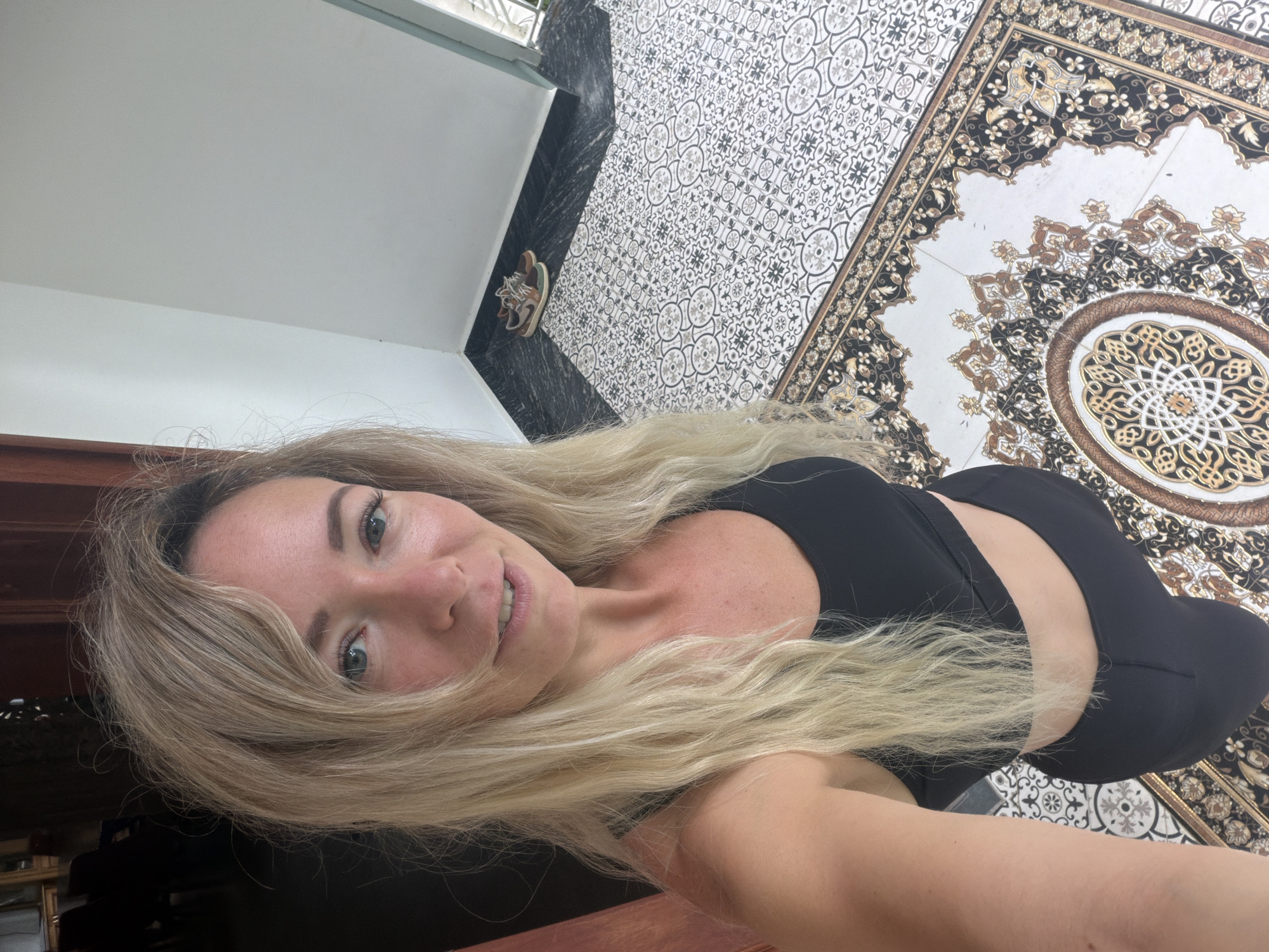A woman with long blonde hair, wearing a black sports bra and black leggings, stands on a patterned tiled floor. She is looking up and smiling slightly at the camera. The floor tiles are black and white with intricate patterns. There is also a section of larger, ornate tiles with brown, gold, and black designs. To the left, a dark wooden door frame is visible. To the right, a white wall meets a dark marble baseboard. In the background, a pair of sneakers rests on the baseboard. The scene appears to be an indoor entryway or hallway. The location is identified as Xuân Trường - Đà Lạt, Vietnam.