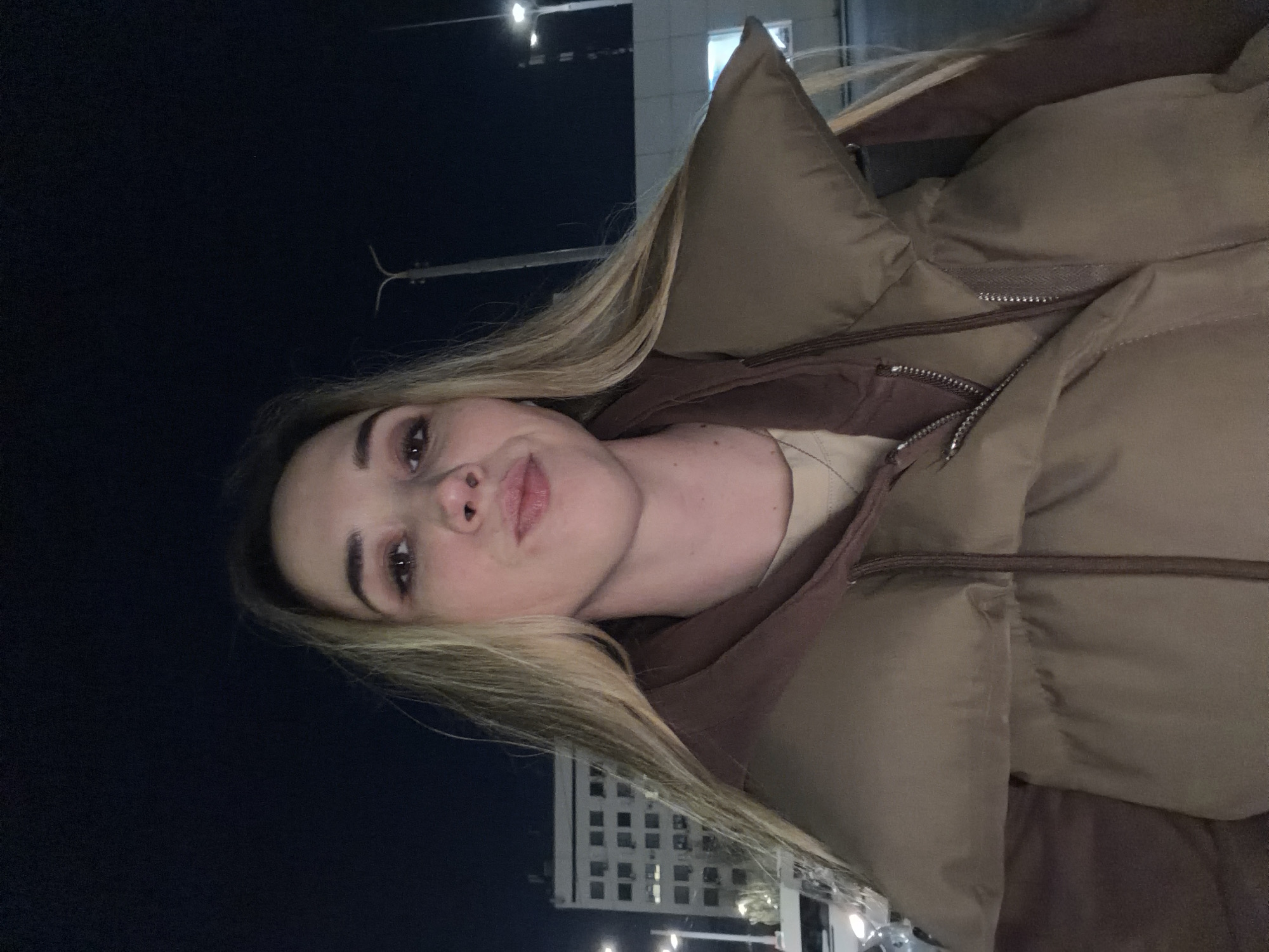 A woman with blonde hair is posing for a selfie at night in Ovoshchnoy, Russia. She is wearing a light brown puffer jacket with a brown hoodie underneath. Behind her, a building with many windows is visible, and there is a dark sky above. Streetlights are also present in the background.