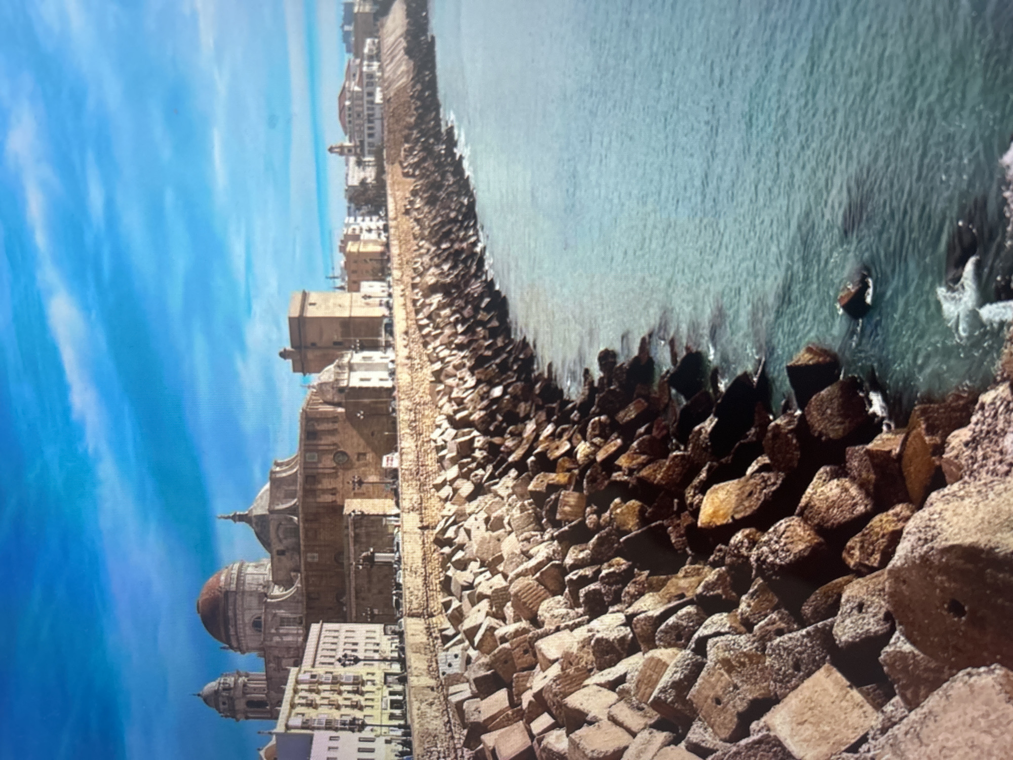 The image displays a panoramic view of a historic coastal city under a bright, clear sky. On the left, a dense cluster of buildings forms the city's skyline, with a grand cathedral being the most prominent structure. The cathedral features a large, distinctive golden-brown dome and multiple towers, showcasing classical European architectural elements. Adjacent to it are various multi-story buildings, some appearing residential with visible windows and balconies.
A substantial breakwater, constructed from numerous large, angular concrete blocks, separates the city from the sea. These irregularly shaped blocks, predominantly in shades of brown and grey, are densely packed and line the entire waterfront, serving as a protective barrier. Many of the blocks show signs of weathering or marine deposits.
To the right, the sea extends, exhibiting vibrant turquoise and deep blue hues. The water surface is textured with small ripples and gentle waves, with white foam visible where the waves meet the rocky breakwater. The sky above is a brilliant, unblemished blue, with a few faint, wispy clouds scattered across it, indicating sunny weather. The overall impression is one of a serene and historic Mediterranean or European coastal city, strongly reminiscent of places like Cadiz, Spain. No people or visible text are present in the image.