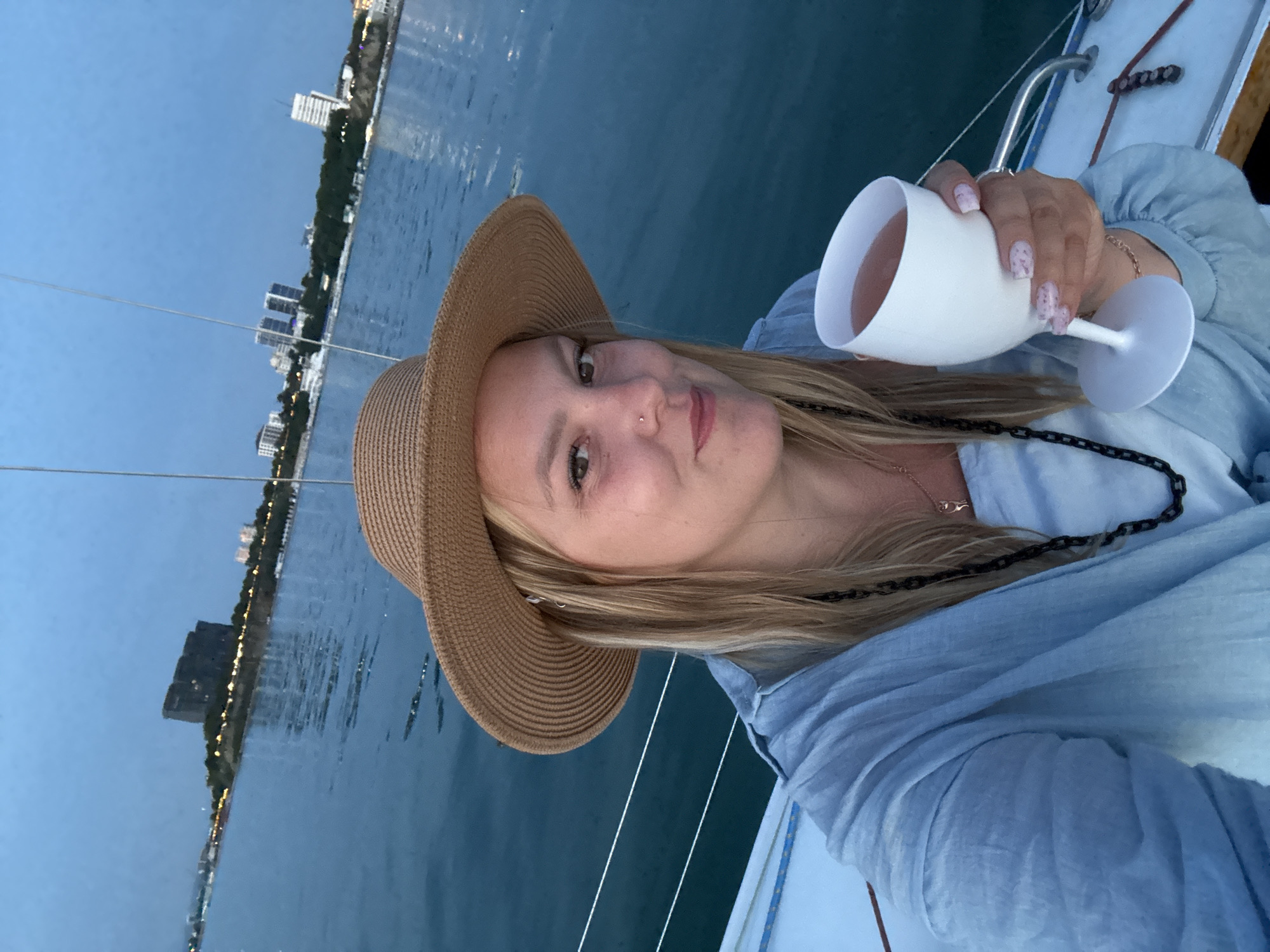 The image depicts a woman on a boat at twilight, situated in what is identified as the city of Krasny, Russia. The woman, with light-colored hair, is wearing a light blue long-sleeved top and a straw hat. She has a small silver nose piercing on her right nostril and a delicate gold-colored necklace, along with a black chain necklace. Her fingernails are manicured with a speckled pink design. She is holding a white, opaque stemmed cup filled with a pink liquid, possibly a beverage like rosé wine. She is looking directly at the viewer with a subtle smile. The foreground shows parts of the white boat deck and railings, with calm, dark blue water surrounding it. In the background, a cityscape is visible along a coastline under a dark blue sky. To the left, a large, dark, multi-story building stands prominently on an elevated landmass or island, displaying some illuminated windows. Further along the coastline to the right, several other multi-story buildings, some appearing residential or commercial, are illuminated, indicating an active urban area.