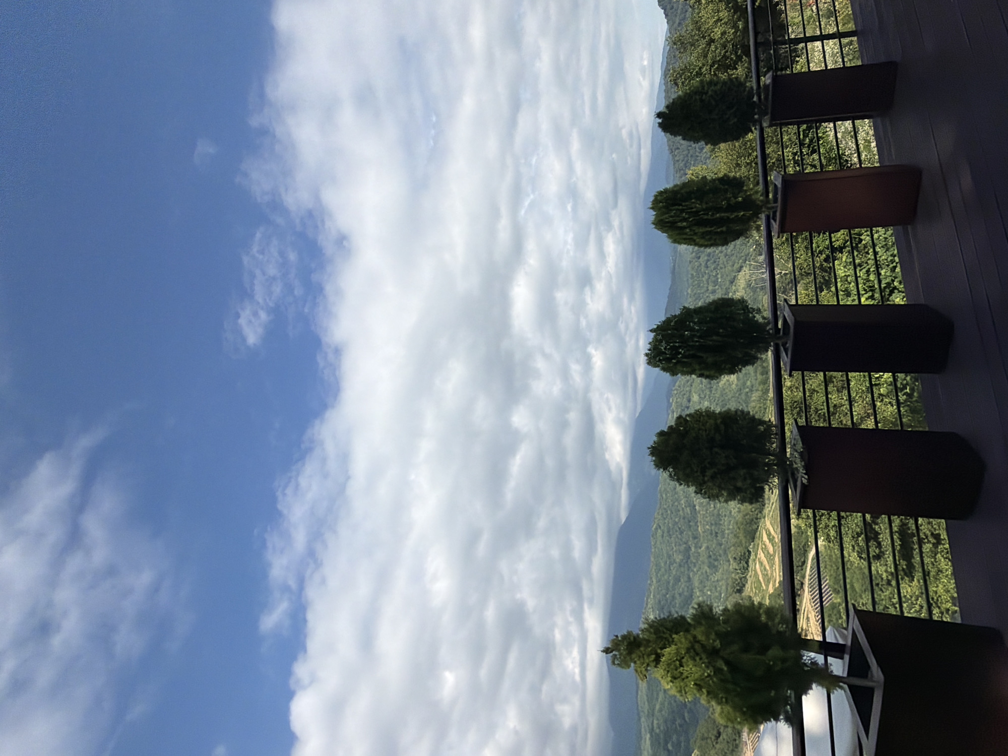 The image shows a view from a balcony in Nizhnyaya Shilovka, Russia. The balcony has a wooden deck and a railing, and several potted evergreen trees are lined up along the railing. Beyond the balcony, a lush green landscape with rolling hills and mountains stretches into the distance under a cloudy blue sky. The overall scene conveys a sense of tranquility and natural beauty.