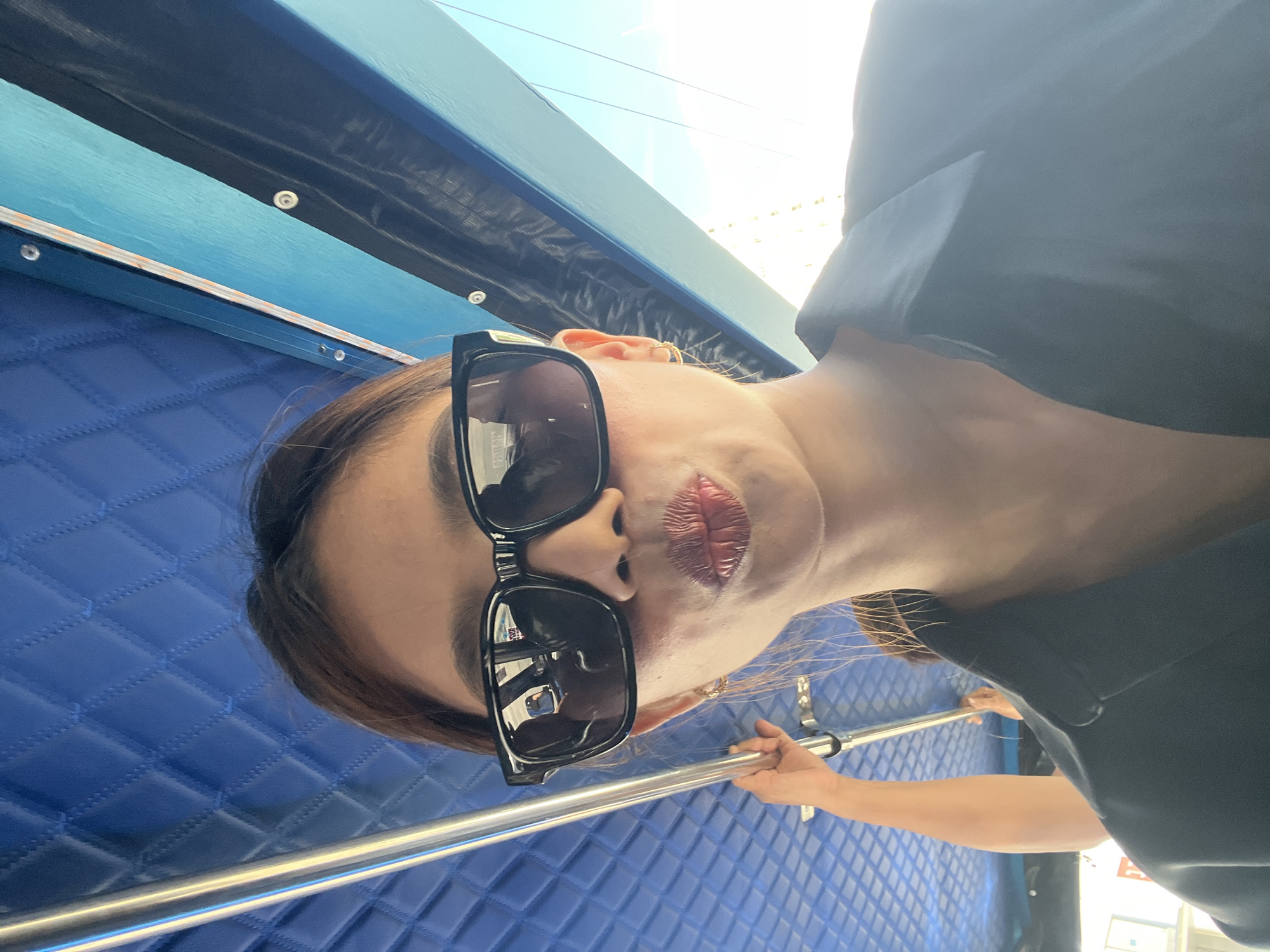 A low-angle, close-up selfie shows a woman wearing black sunglasses and a black collared shirt. She is leaning forward, puckering her lips, and holding a metal pole with her left hand. The background consists of a blue quilted ceiling and bright sunlight. The scene appears to be inside a vehicle, possibly a bus or a boat, given the handrail and the overall structure visible. Without further context, a specific location within Davao City cannot be determined.