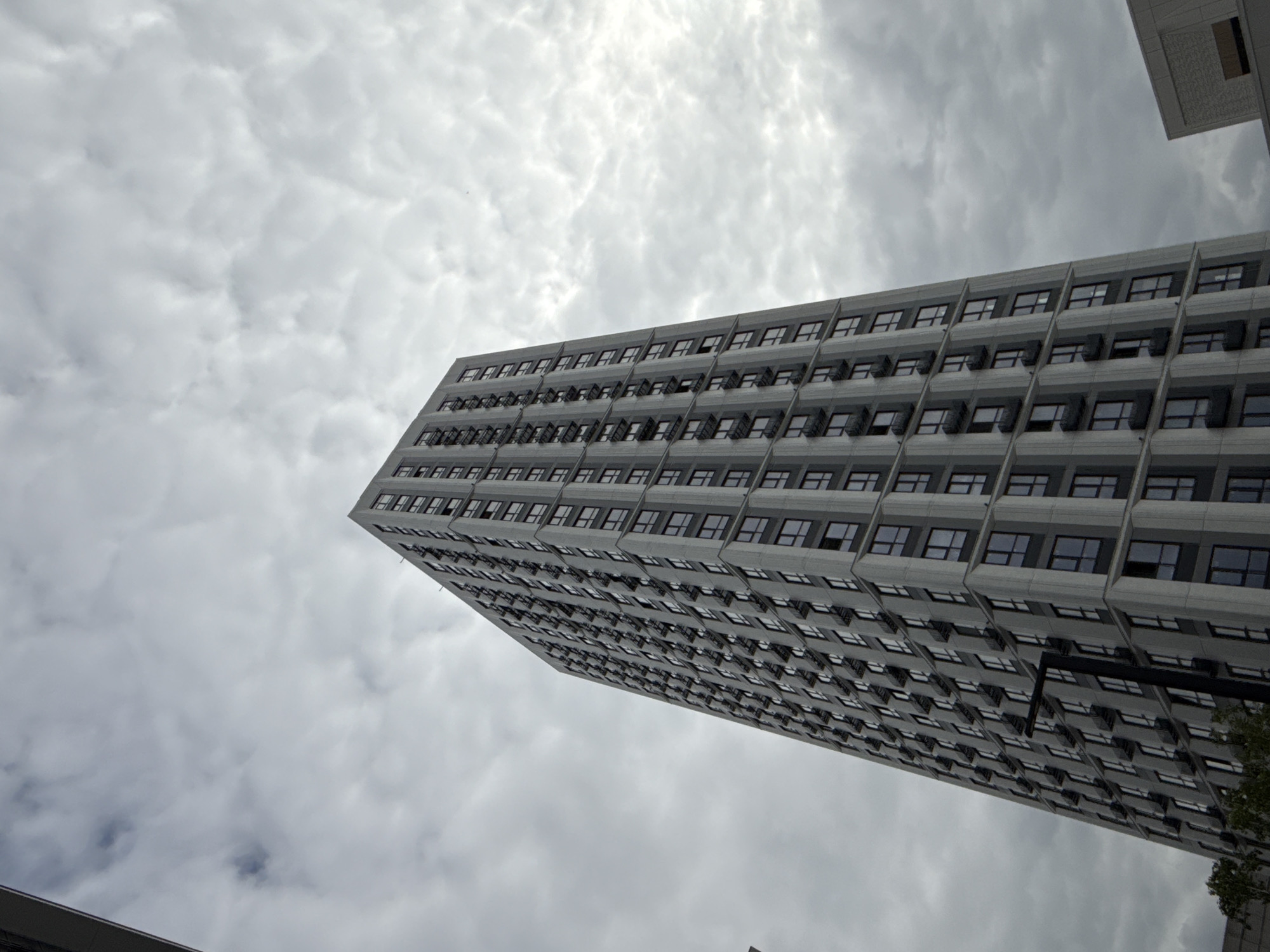 An upward-angled shot captures a towering, modern building against an overcast sky in Moscow, Russia. The building, rendered in shades of gray, occupies the majority of the frame, stretching diagonally from the lower left towards the upper right. Its facade is characterized by a highly repetitive pattern of dark, rectangular windows set within a grid of lighter gray concrete or stone panels. Many of the windows appear slightly ajar or open, suggesting a functional ventilation design.
The sky is filled with dense, textured gray clouds, creating an overall muted and diffused light, indicative of an overcast day. A bright spot in the upper central sky suggests where the sun might be, though it is fully obscured by clouds. In the upper right corner, a small section of another building or structure, also gray, is visible, adding to the urban context. A hint of green foliage, possibly from a tree, can be seen at the bottom right edge of the frame, contrasting with the dominant architectural and sky elements. The low perspective emphasizes the building's height and imposing presence. There is no visible text or discernible activity in the static scene.