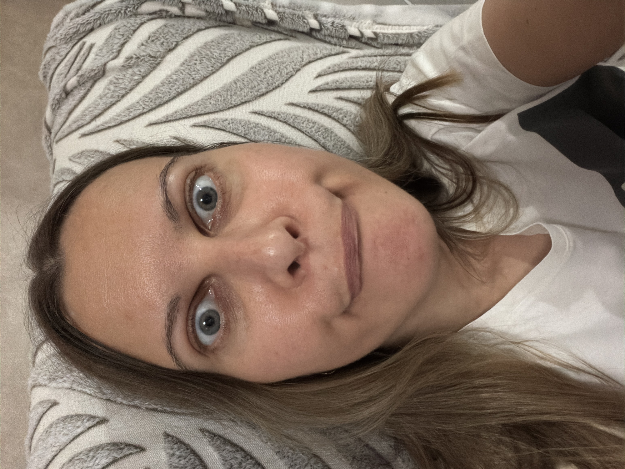 A close-up, high-angle, selfie-style shot captures a woman with light brown hair and blue eyes. Her expression is a wide-eyed, slightly puckered pout. She is wearing a white t-shirt with a dark grey graphic on the left side. The background consists of a textured pillow with a leaf-like pattern in shades of grey and white, and a neutral-colored wall. The scene appears to be indoors, possibly a living room or bedroom, in Izhevsk, Russia.