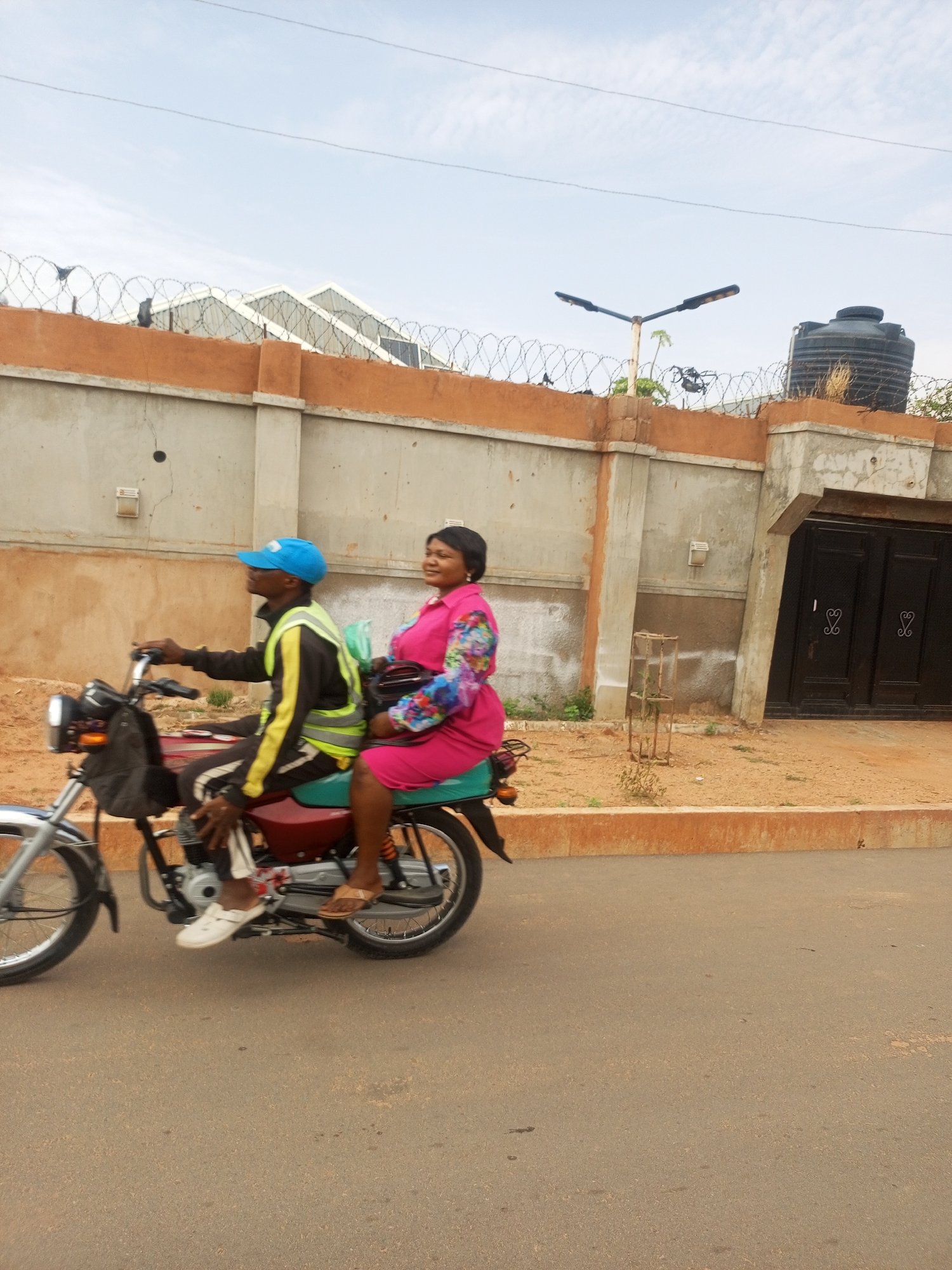 A man and a woman are riding on a motorcycle along a street in Tempure, Nigeria. The man, driving the motorcycle, wears a blue cap, a black jacket with yellow stripes, white sneakers, and dark pants. He has a black bag strapped to the front of the motorcycle. The woman, seated behind him, wears a pink dress with a floral pattern on the sleeves and brown sandals. She is holding a bag. The motorcycle is red and silver.
They are riding on an asphalt road. A wall lines the side of the road, topped with barbed wire. Behind the wall are buildings, a water tank, and a street light. The sky is pale blue.